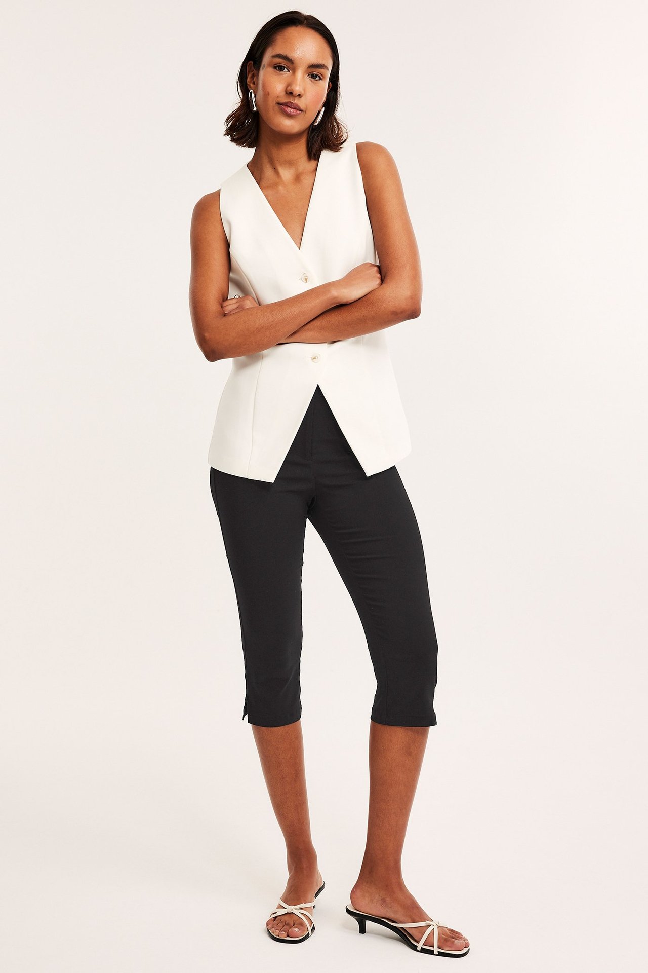 Edestäpäin: nainen mustissa bengaliini caprihousuissa, valkoisessa hihattomassa liivissä ja valkoisissa sandaaleissa.