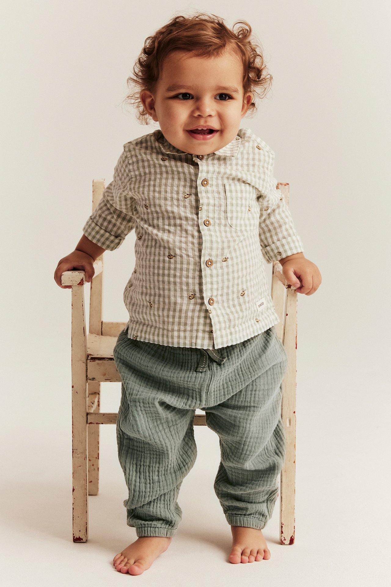 Frontutsikt: baby i grønne musselinbukser og en lysrutet skjorte med ankertrykk, holder stol.