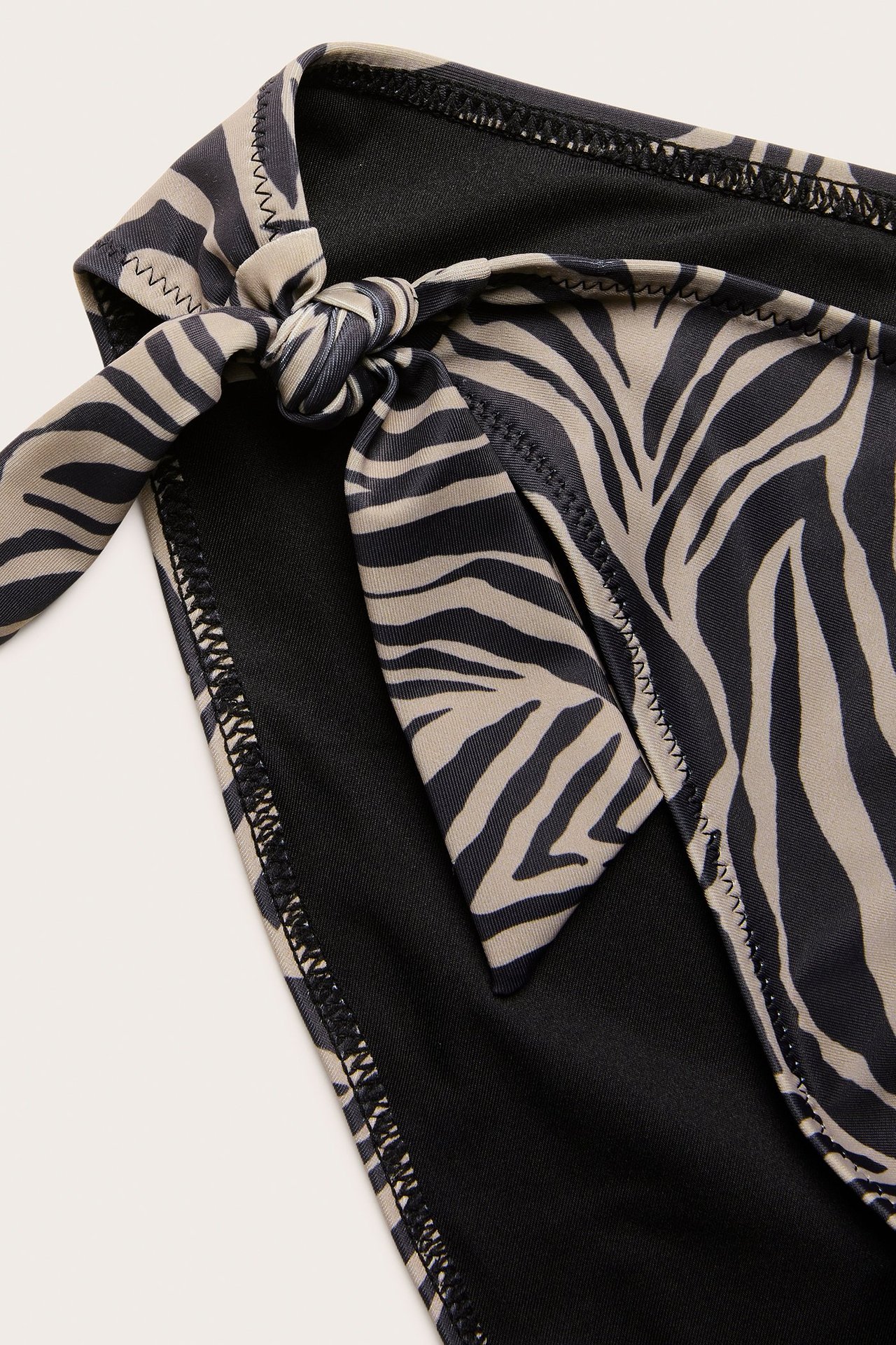 Zbliżenie na ciemnobeżowe i czarne figi bikini w zebrę z ozdobnymi wiązaniami po bokach.