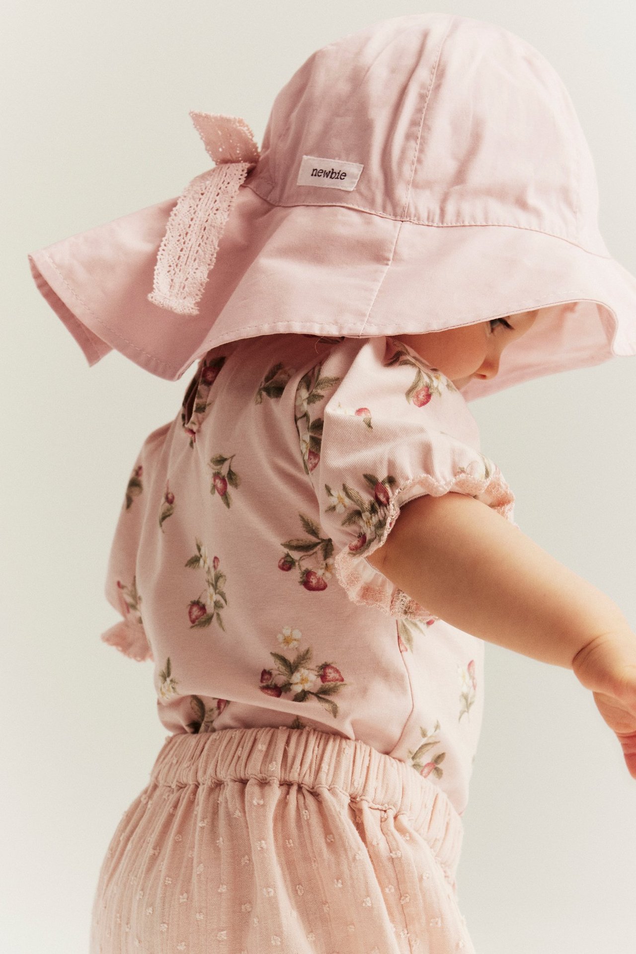 Sivukuva vauvasta yllään vaaleanpunainen aurinkohattu pitsirusetilla, mansikkakuvioinen toppi ja vaaleanpunaiset kuvioidut shortsit.