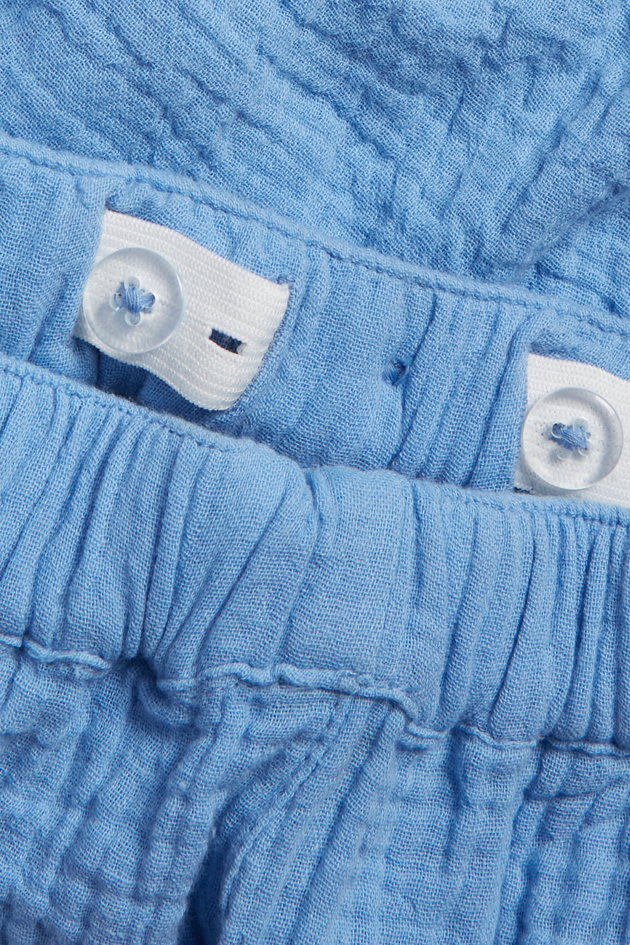 Närbild av blå muslinshorts med justerbar resår i midjan och genomskinliga knappar.