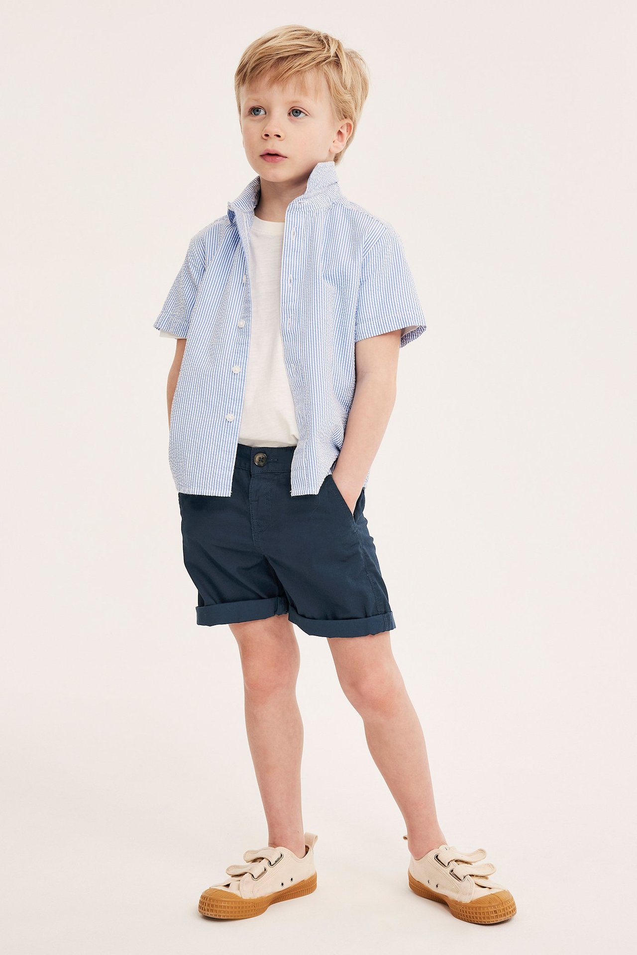 Edestä näkymä lapsesta tummansinisissä puuvillashortseissa, vaaleansinisessä raidallisessa paidassa ja valkoisessa t-paidassa, sekä beigessä tennareissa.