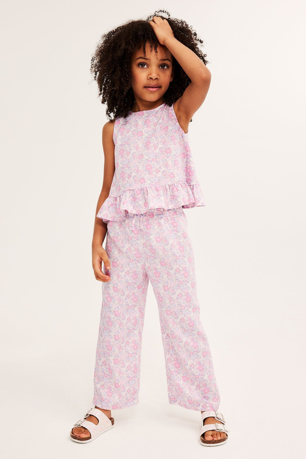 Framifrån: Ett barn i rosa blommiga culottebyxor i viskos, matchande ärmlös topp och vita sandaler.