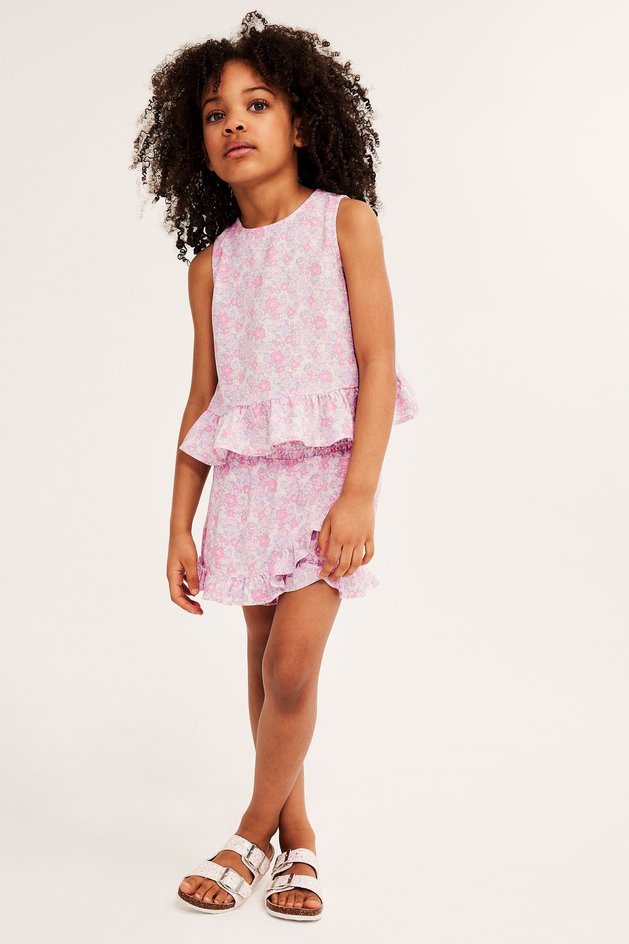 Widok z przodu dziecka w różowej, kwiecistej spódnico-spodenkach z wiskozy, pasującym topie i srebrnych sandałach.