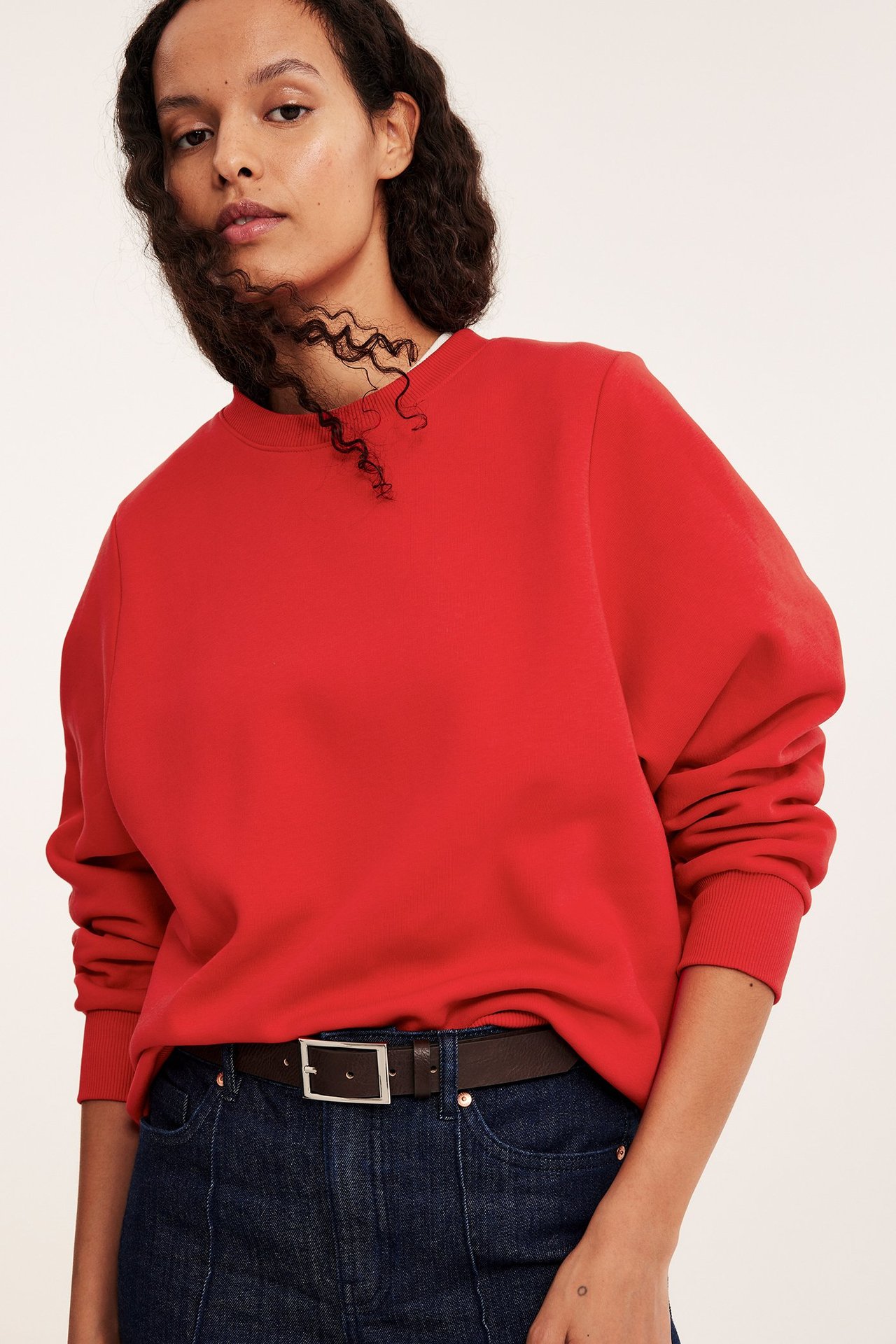Widok z przodu: kobieta w czerwonej bluzie z mieszanki bawełny, ciemnoniebieskich dżinsach i brązowym pasku.