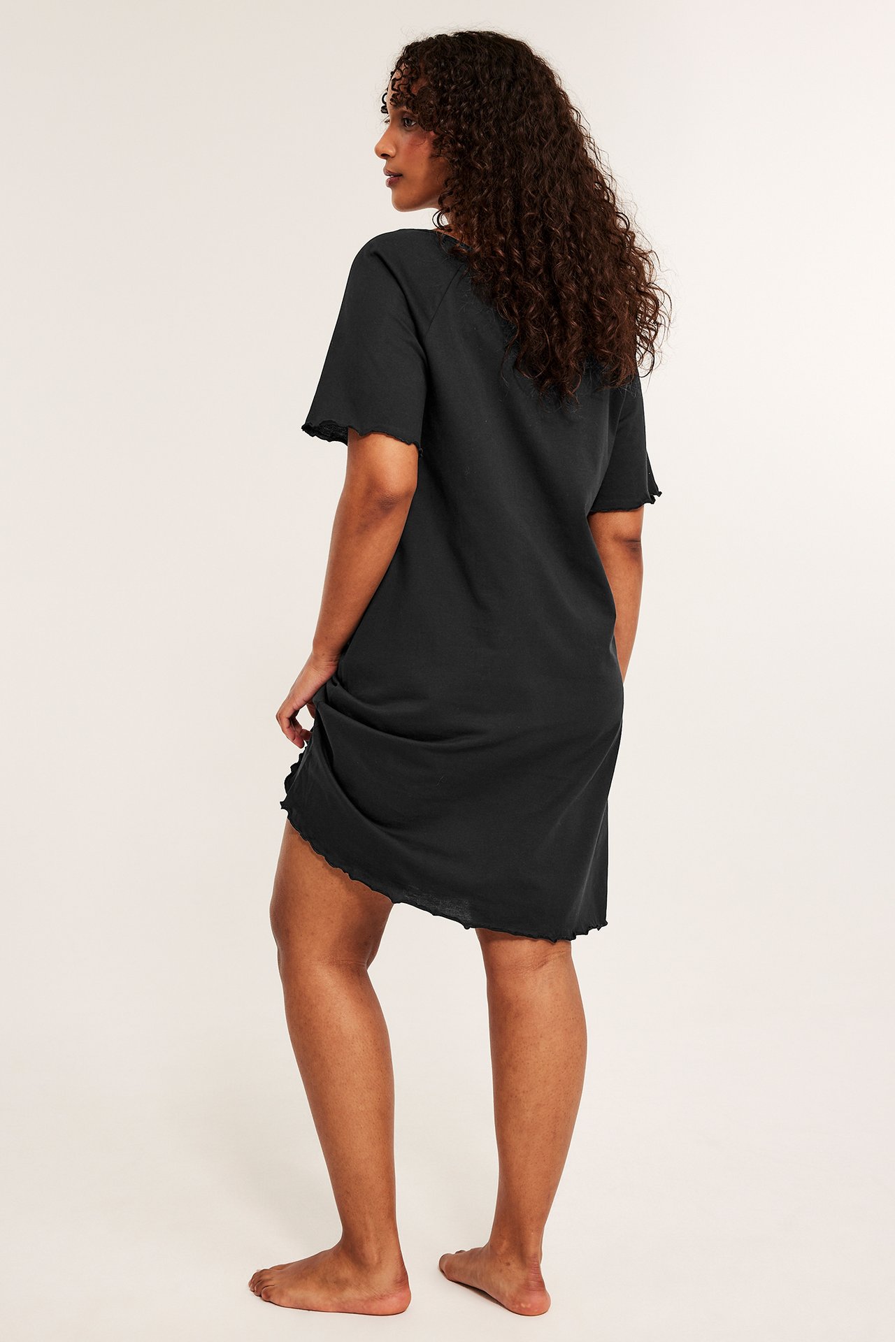 Sidevisning av en kvinne i en løstsittende svart bomullsnattkjole med korte ermer og bølgete kanter.