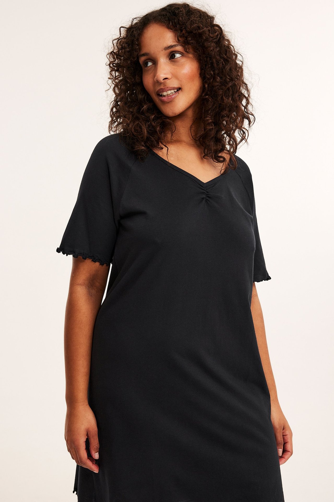 Edestäpäin, nainen yllään musta puuvillatrikoo yöpaita V-pääntiellä, rypytyksellä ja aaltoilevilla lyhyillä hihoilla.