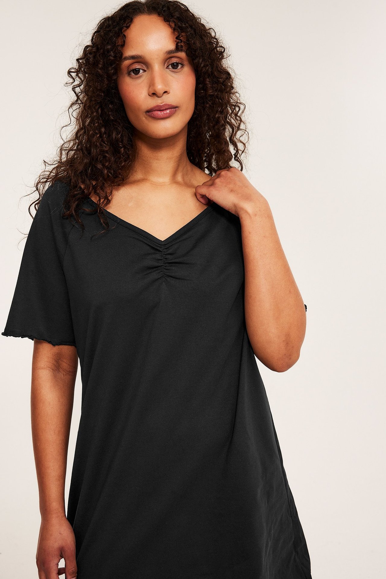 Edestäpäin, nainen mustassa puuvillayöpaidassa, jossa on rypytetty V-pääntie ja aaltoilevat lyhyet hihat.