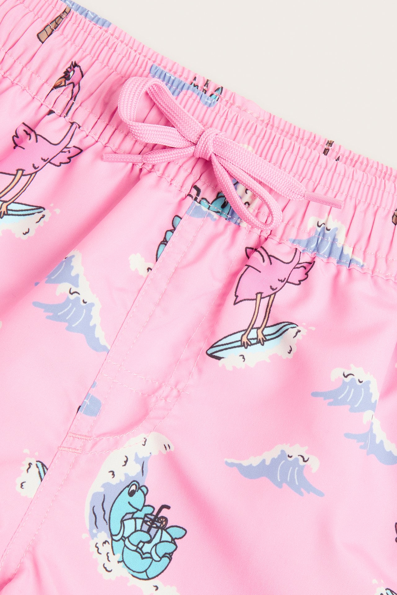 Närbild på ljusrosa badshorts för barn med surfande flamingos och sköldpaddor, resår i midjan och rosa dragsko.