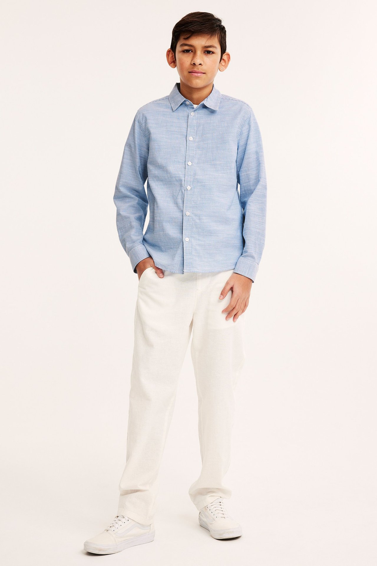Widok z przodu dziecka w jasnoniebieskiej koszuli w paski, białych spodniach i białych trampkach.