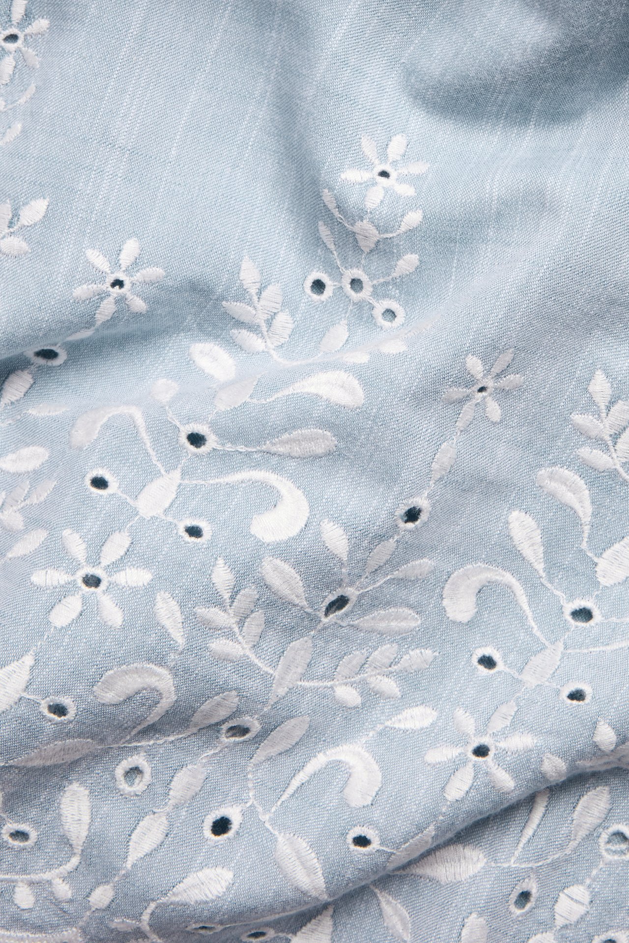 Nærbilde av lyseblått bomulls-chambray-stoff med hvite blomsterbroderier og utskårne detaljer.