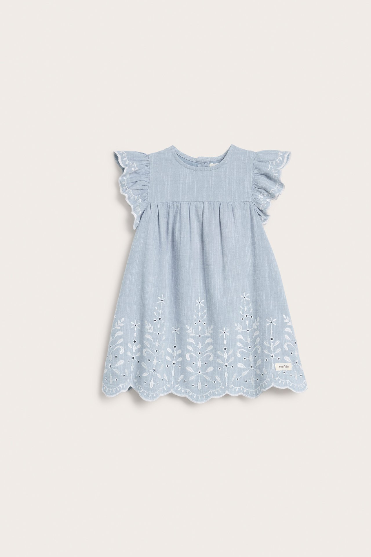 Frontvisning av en lyseblå chambray barnekjole med hvitt broderi, volangermer og knapper bak.
