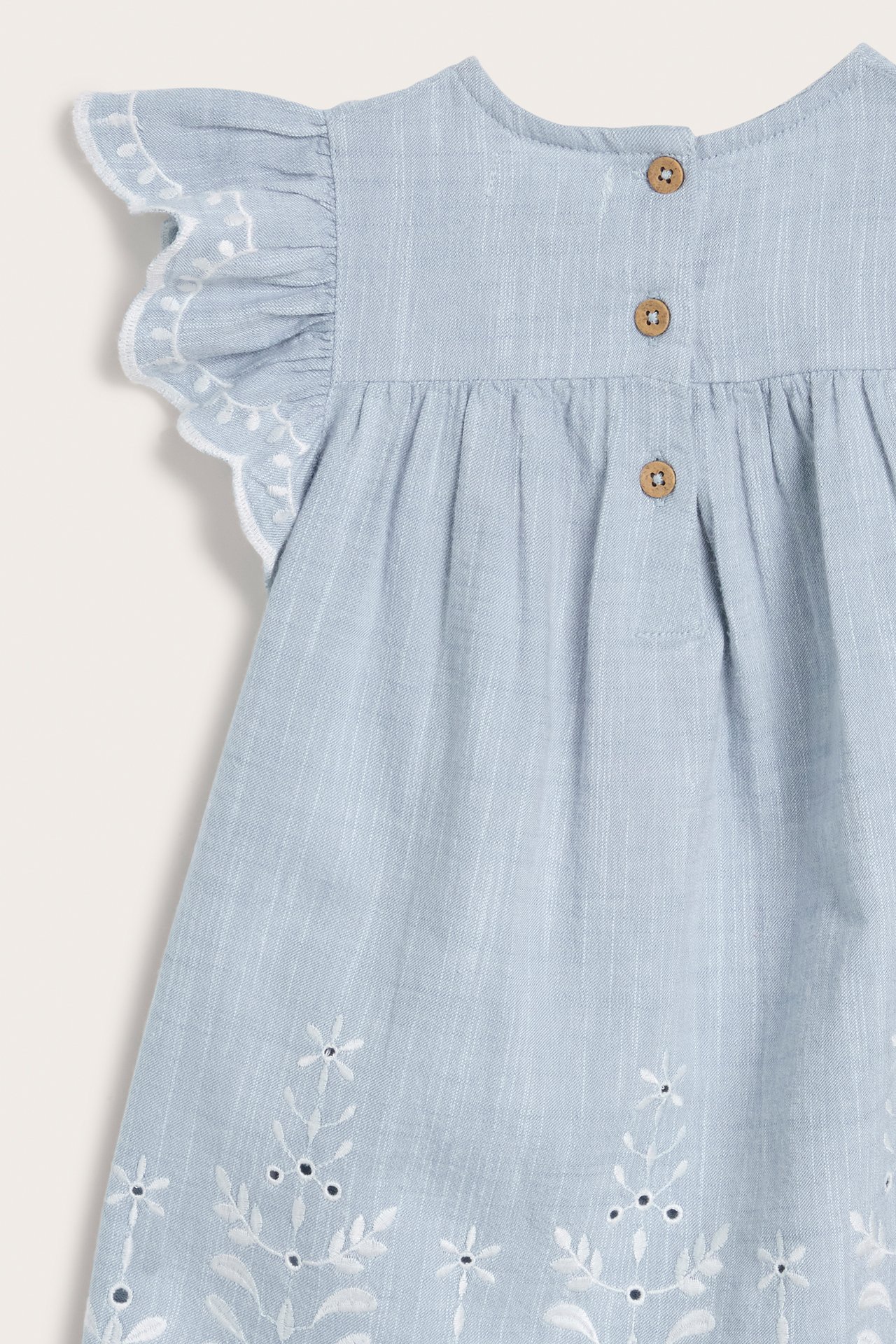 Nærbilde bakfra: lyseblå chambray barnekjole med hvite broderier, rysjeermer og knapper bak.