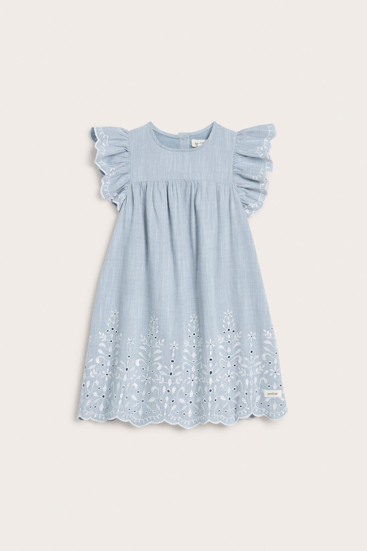 Frontvisning av en lyseblå chambray barnekjole med hvite broderier og volangermer.