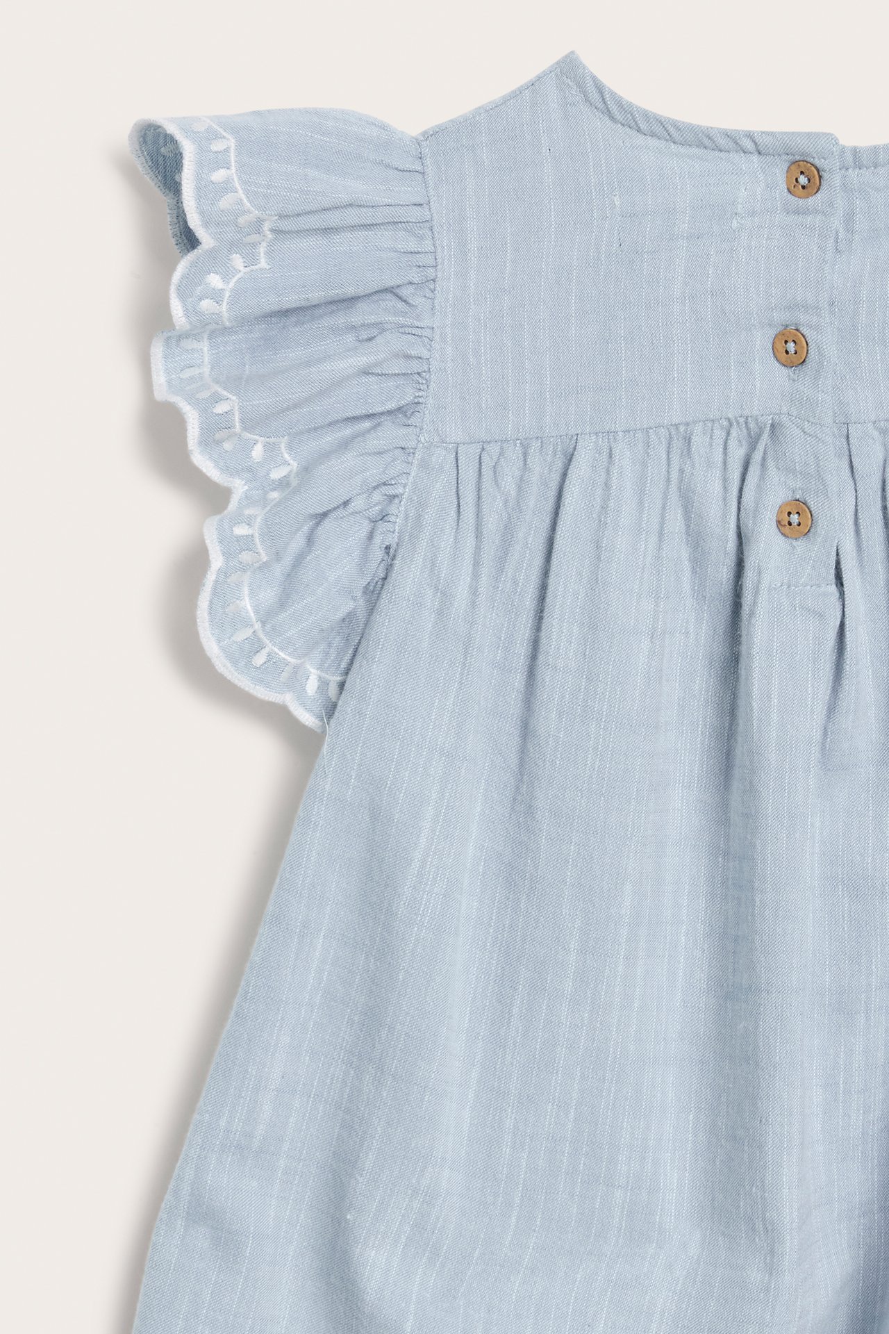 Nærbilde bakfra av en lyseblå chambray barnekjole med hvite broderte volangermer og treknapper.
