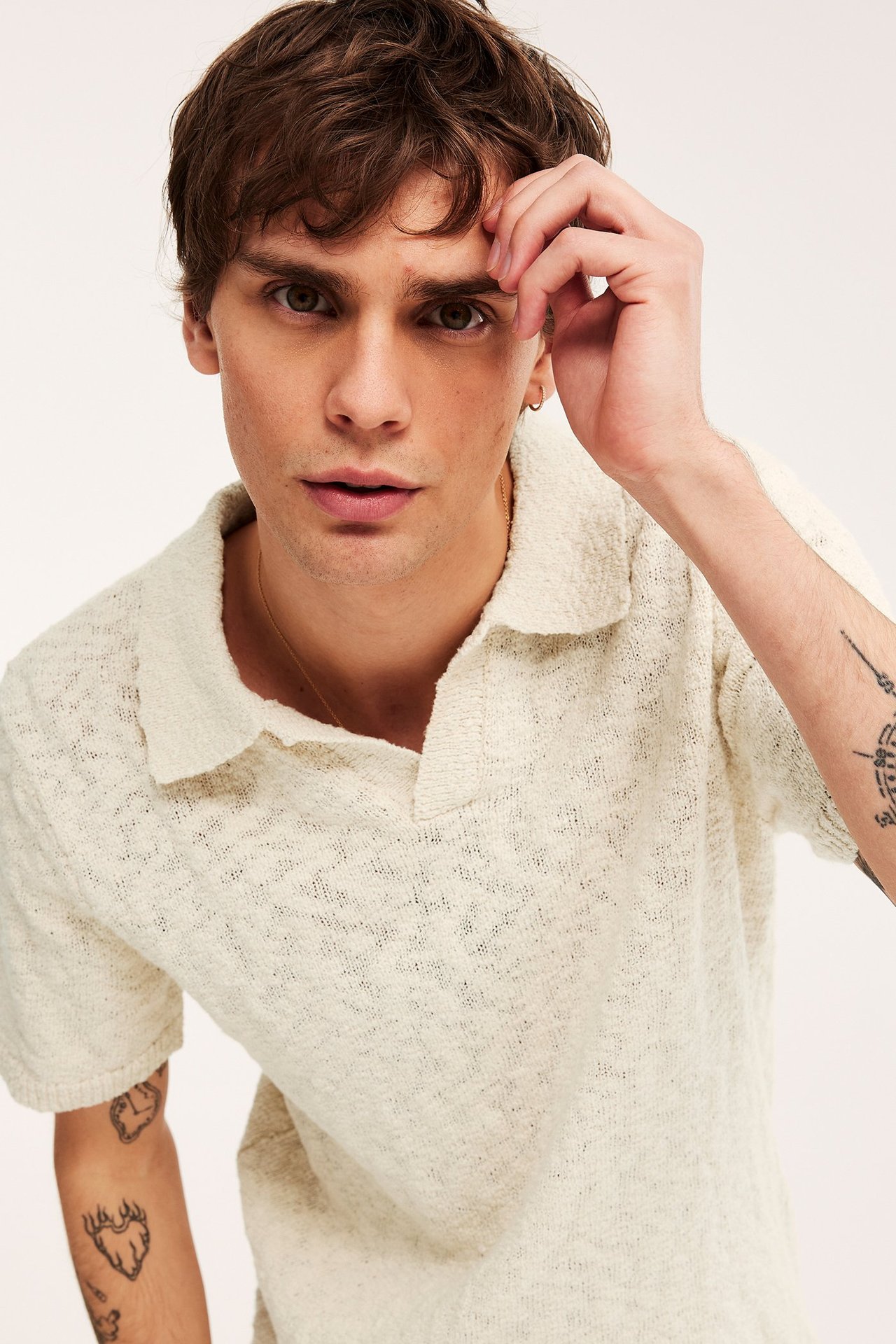 Lähikuva: mies yllään luonnonvalkoinen struktuurineulottu lyhythihainen pikeepaita, kultaketju ja korvakoru.