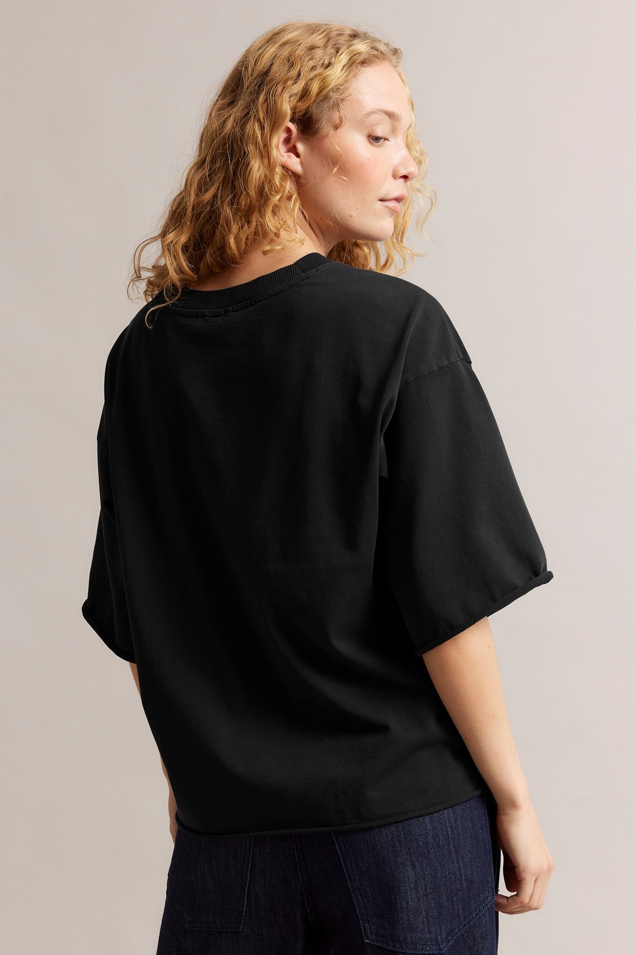 Ryggvy av kvinna i löst sittande svart tvättad bomulls-T-shirt med råa kanter och mörkblå jeans.