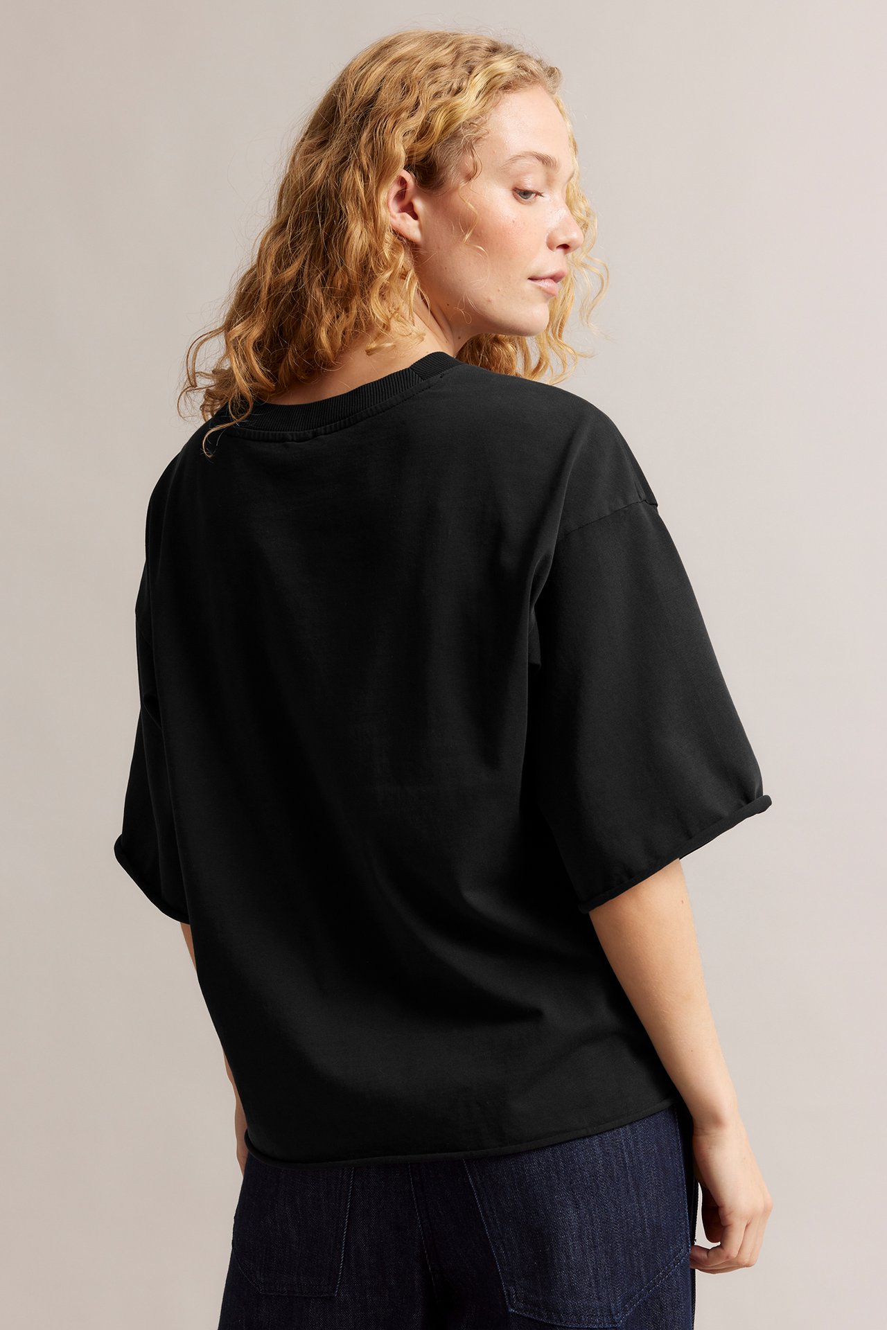 Ryggvy av kvinna i löst sittande svart tvättad bomulls-T-shirt med råa kanter och mörkblå jeans.