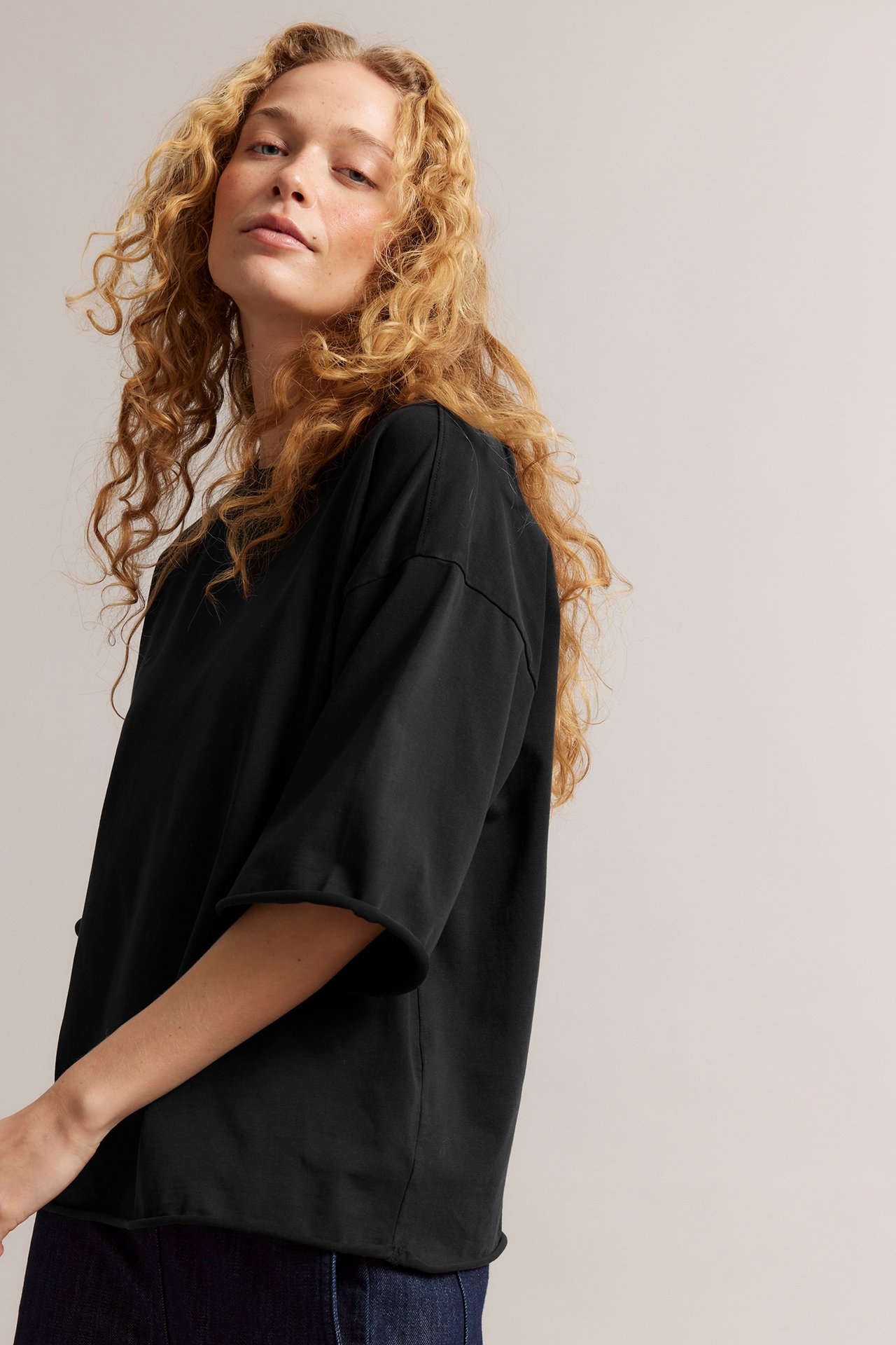 Nærbilde forfra av en kvinne i en svart, vasket, oversized bomulls-t-skjorte med rå kanter over mørke jeans.