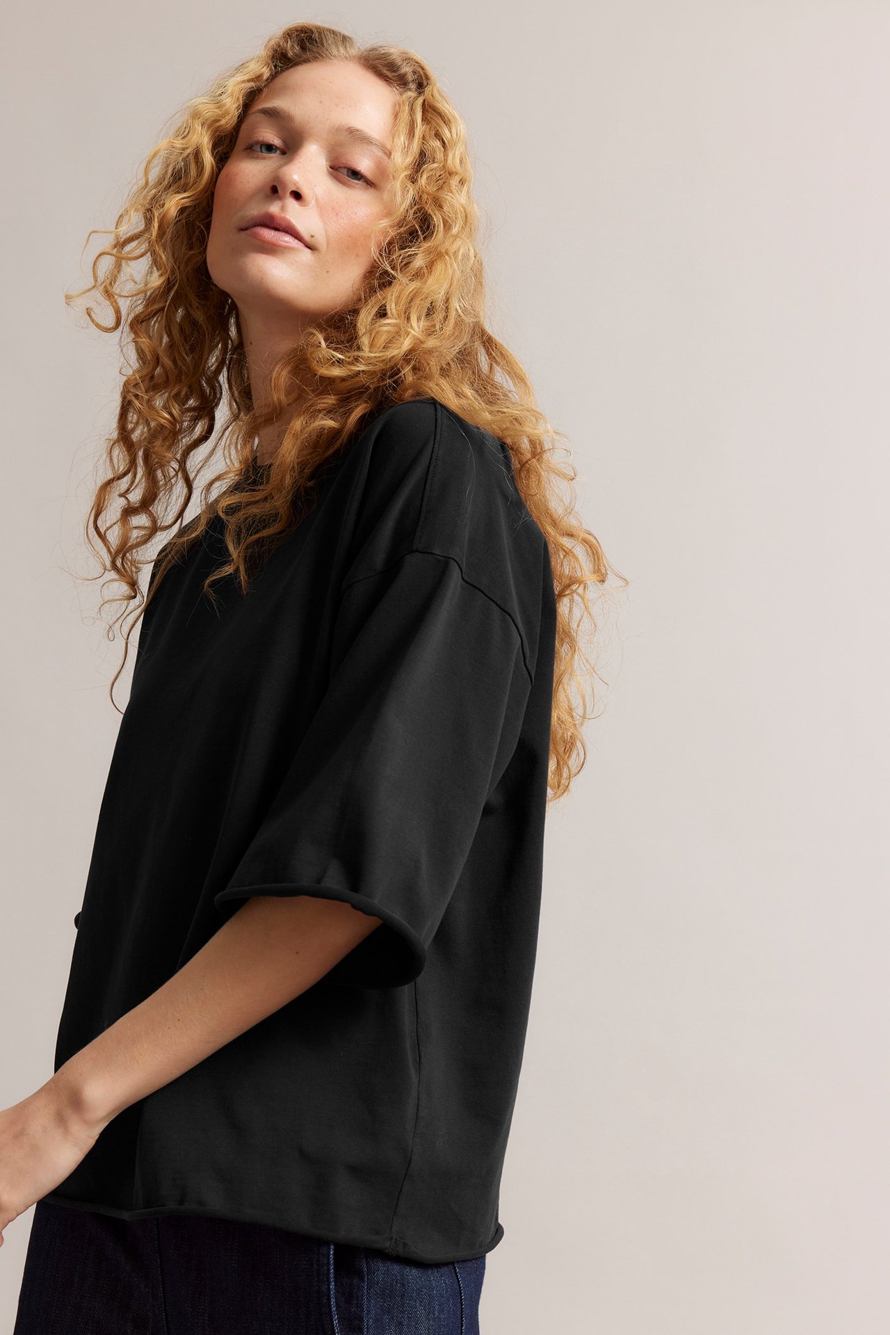 Närbild framifrån på en kvinna i svart, tvättad, oversized bomulls-t-shirt med råa fållar över mörka jeans.