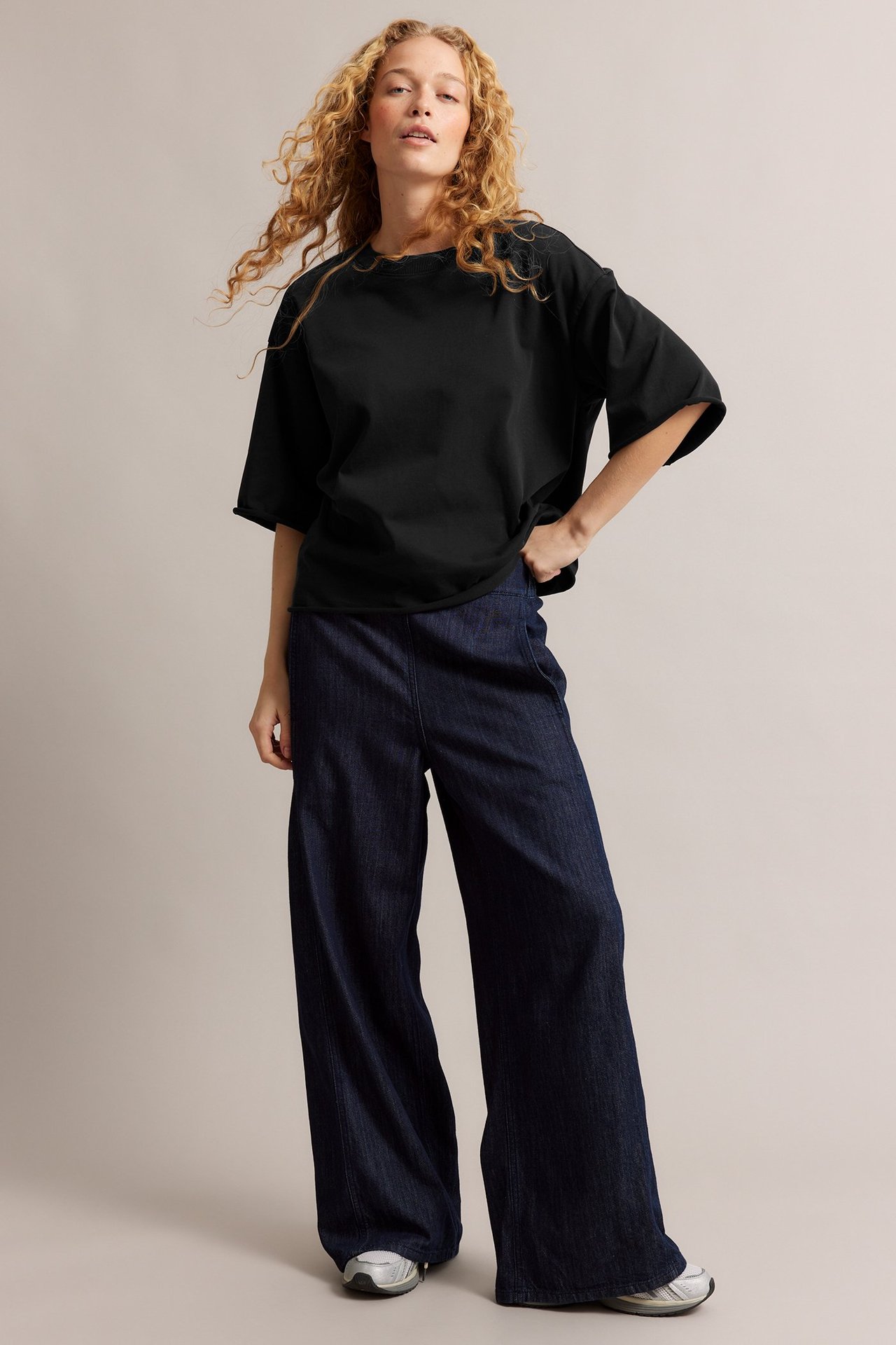 Frontutsikt av en kvinne i svart oversized T-skjorte med råkanter, mørkeblå vide jeans og joggesko.