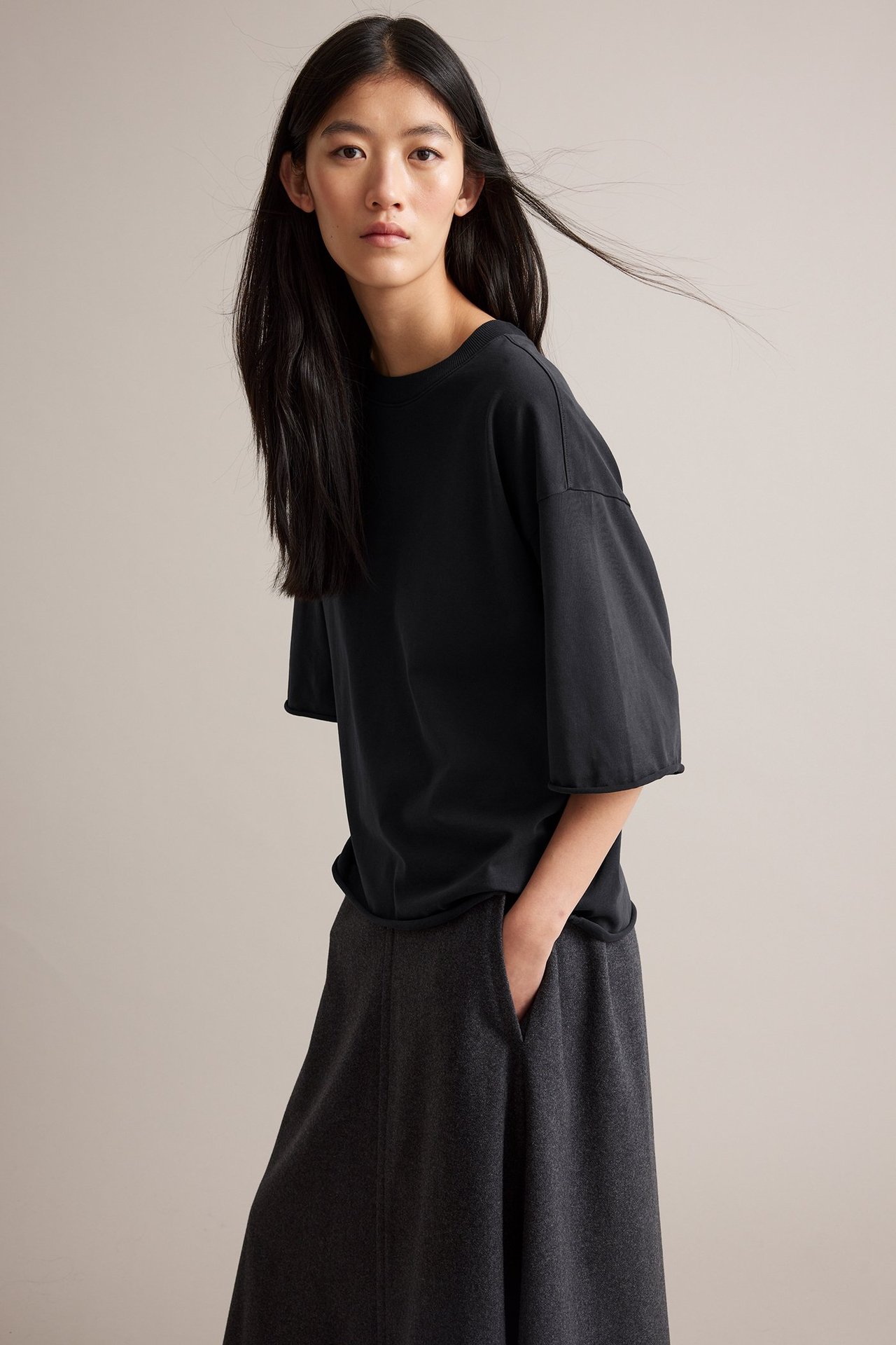 Sida framifrån: kvinna i svart bomulls-t-shirt med råa fållar och mörkgrå lösa byxor.