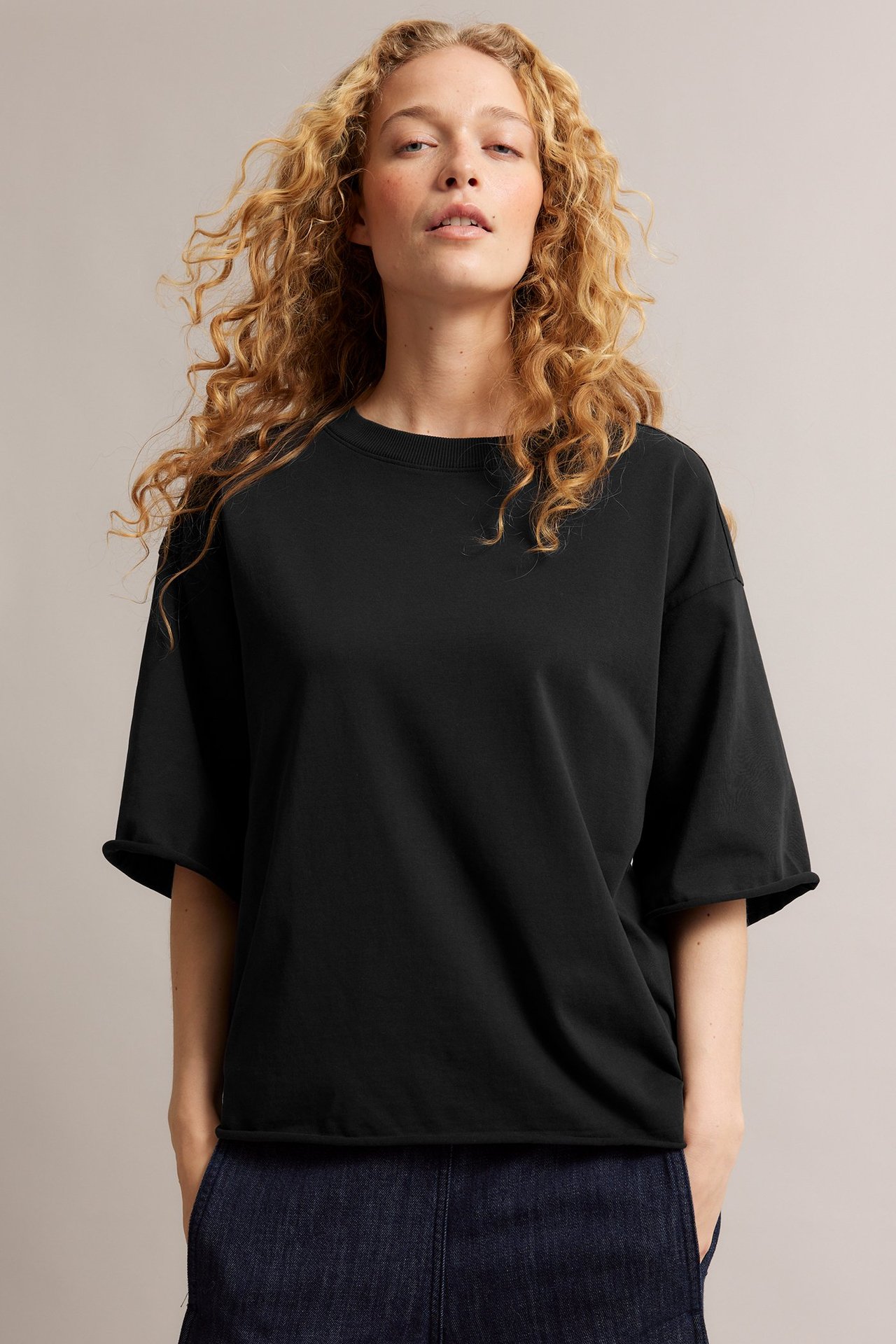 Framifrån, kvinna i en löst sittande svart bomulls-t-shirt med råa fållar och mörkblå byxor.