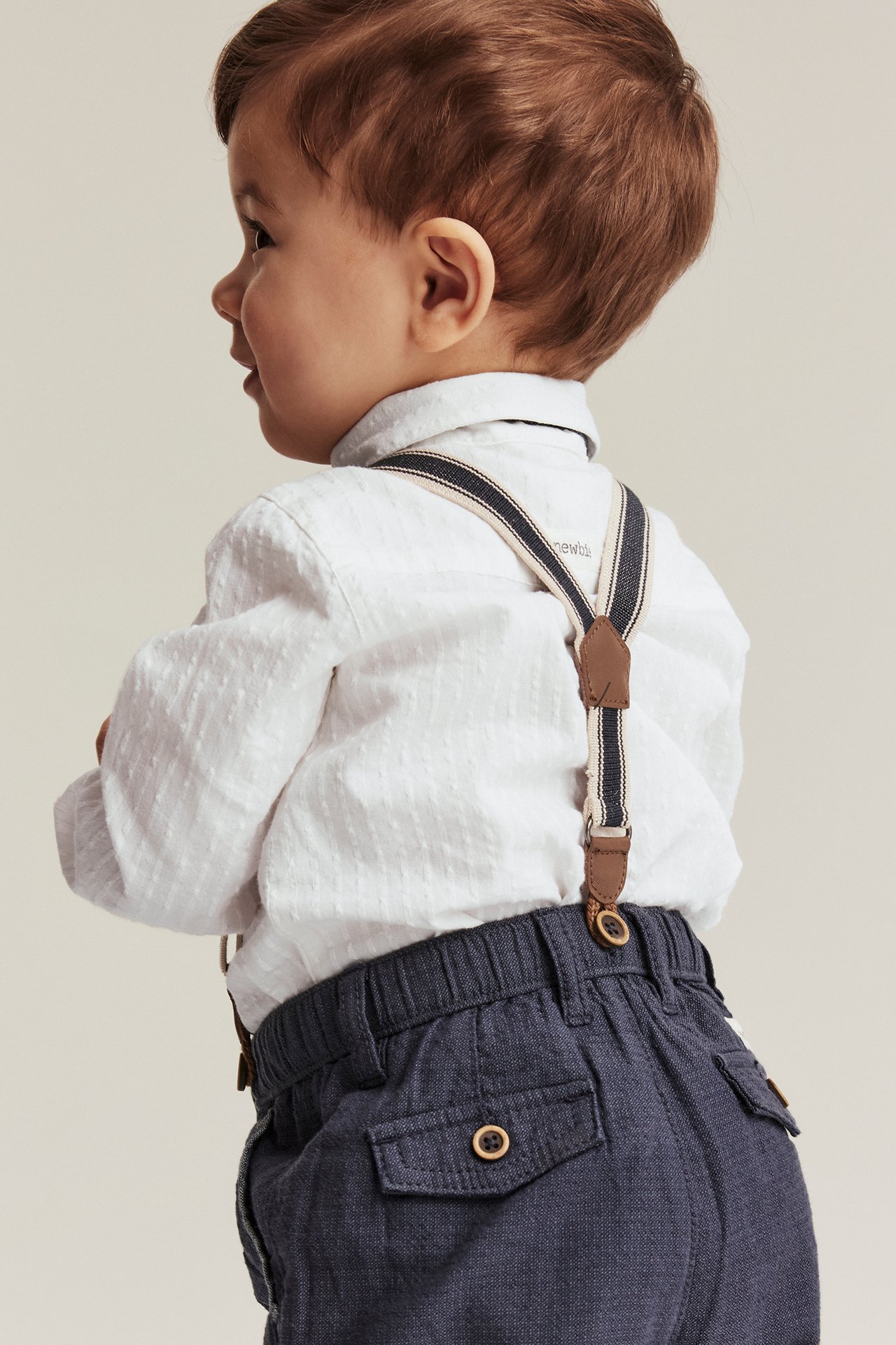 Sidovy av en baby som bär en offwhite texturerad långärmad skjortbody, mörkblå byxor och randiga hängslen.