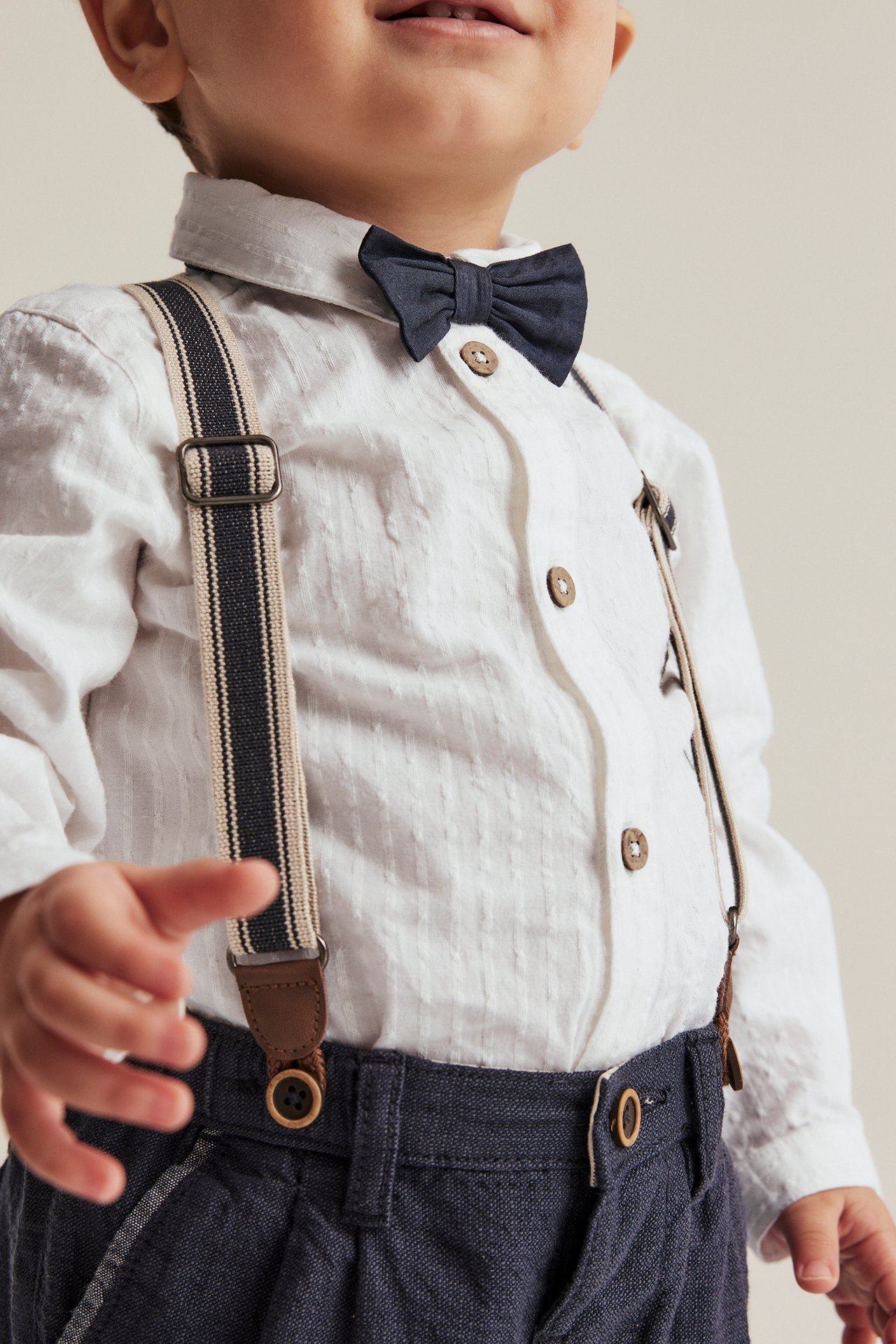 Frontvy: baby i offwhite vävd skjortbody, mörkblå fluga, byxor och randiga hängslen.