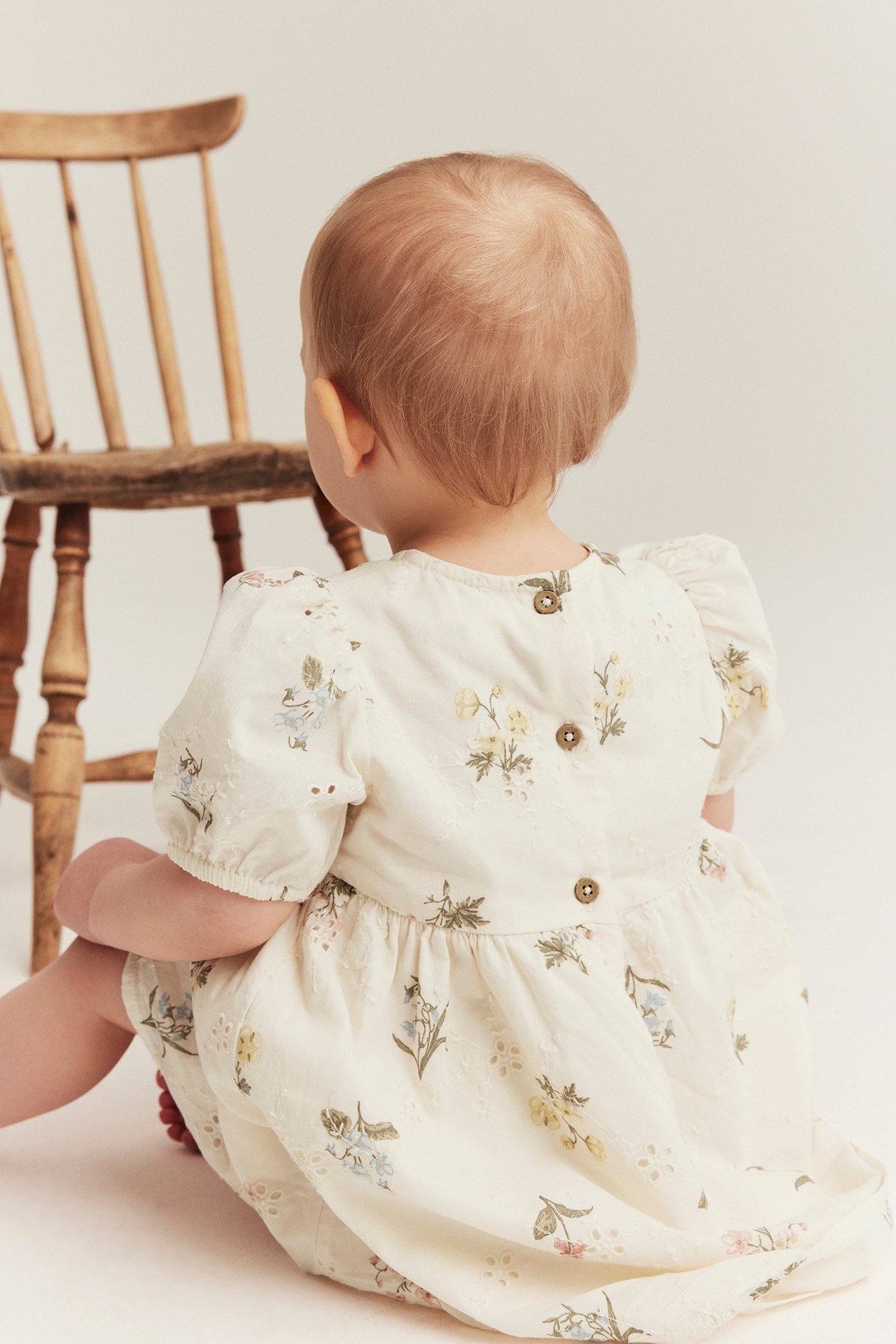 Vauvan selkäkuva offwhite broderie anglaise -mekossa, jossa kukkakuvio ja napit takana.