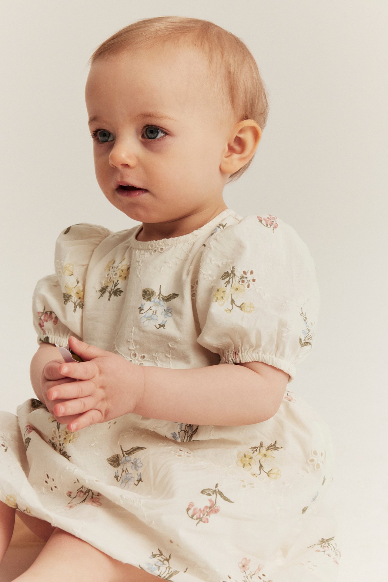 Vauva edestäpäin, yllään luonnonvalkoinen broderie anglaise -mekko kukkakuviolla ja pussihihoilla.