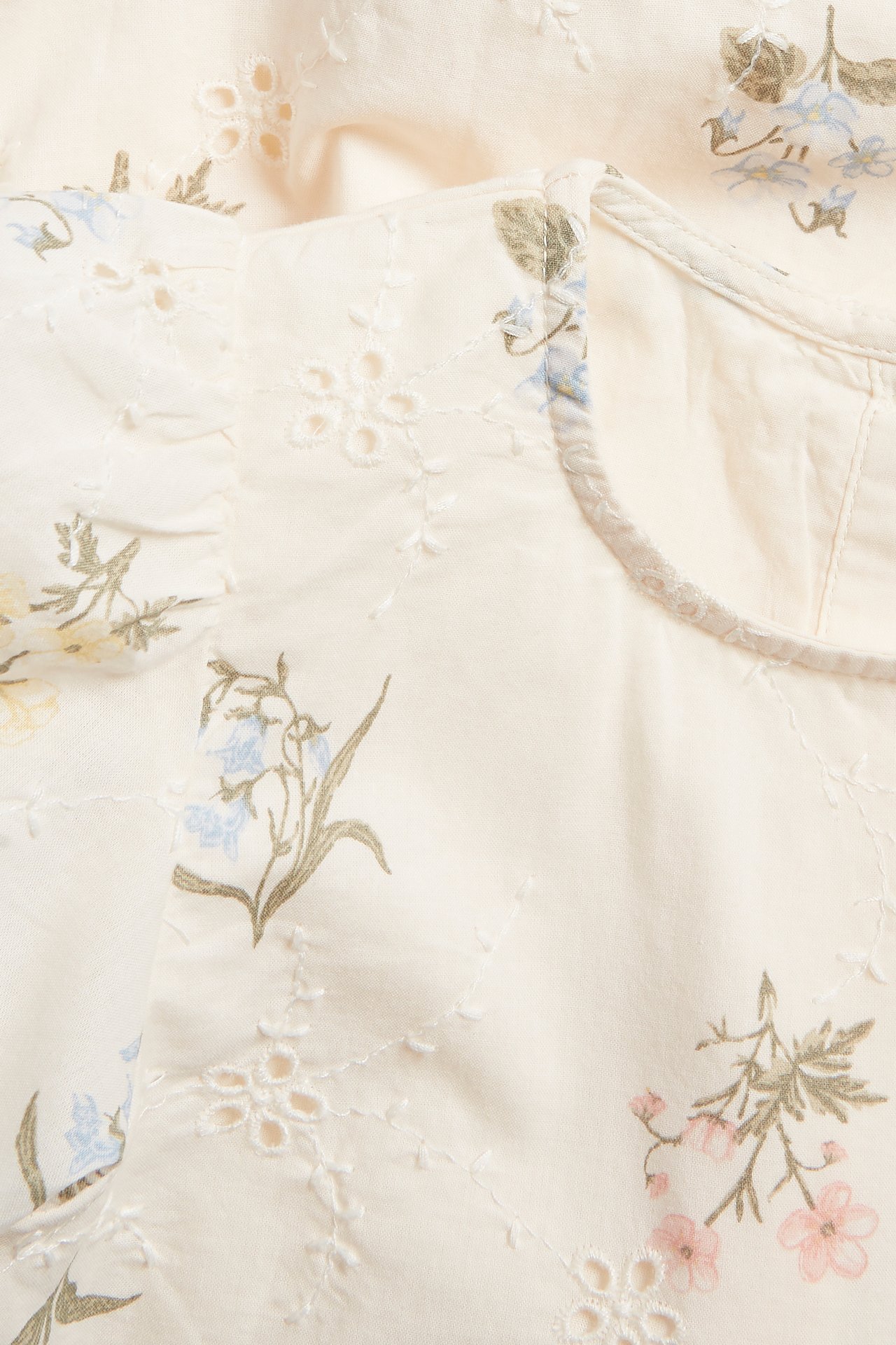 Lähikuva luonnonvalkoisesta kudotusta mekosta, jossa broderie anglaise ja painettuja sinisiä, vaaleanpunaisia ja vihreitä kukkia.