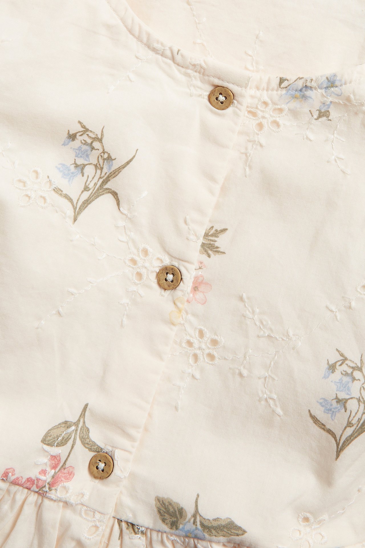 Lähikuva luonnonvalkoisesta mekosta, jossa broderie anglaise, painetut siniset ja vaaleanpunaiset kukat, ruskeat napit.
