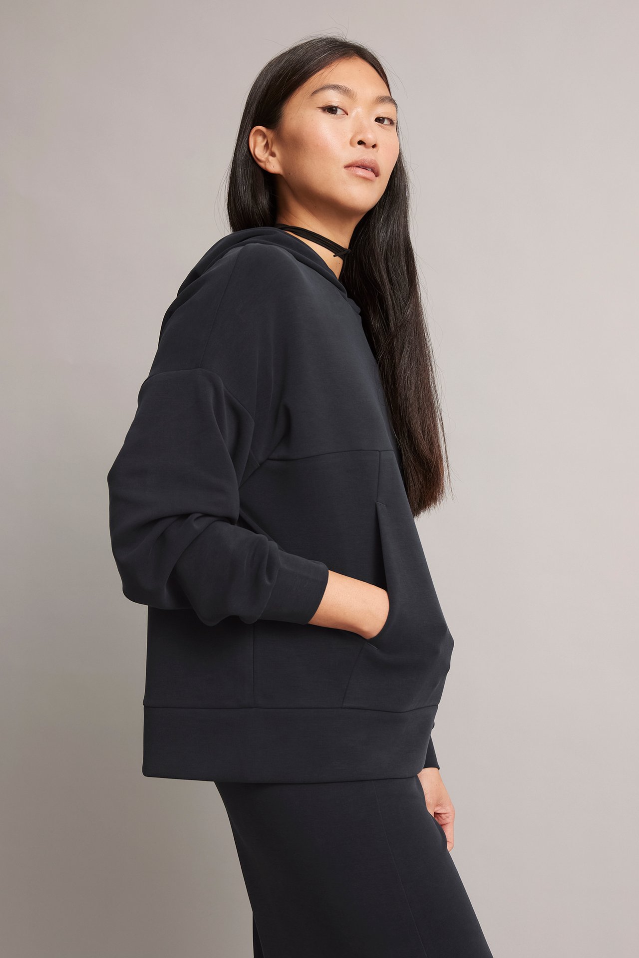 Sivukuva naisesta mustassa Tencel/modaali hupparissa, jossa kengurutasku, musta hame ja choker.