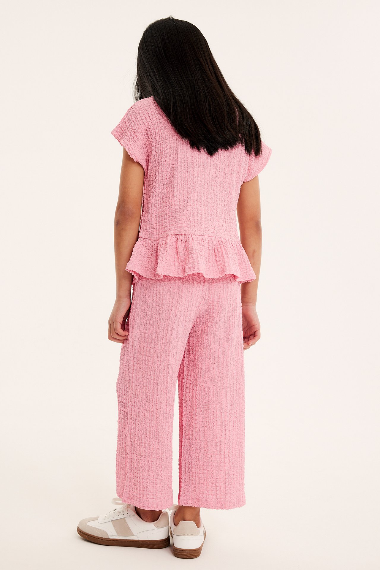 Bakifrån, ett barn i rosa strukturerade culottebyxor, matchande peplumtopp och vita sneakers.