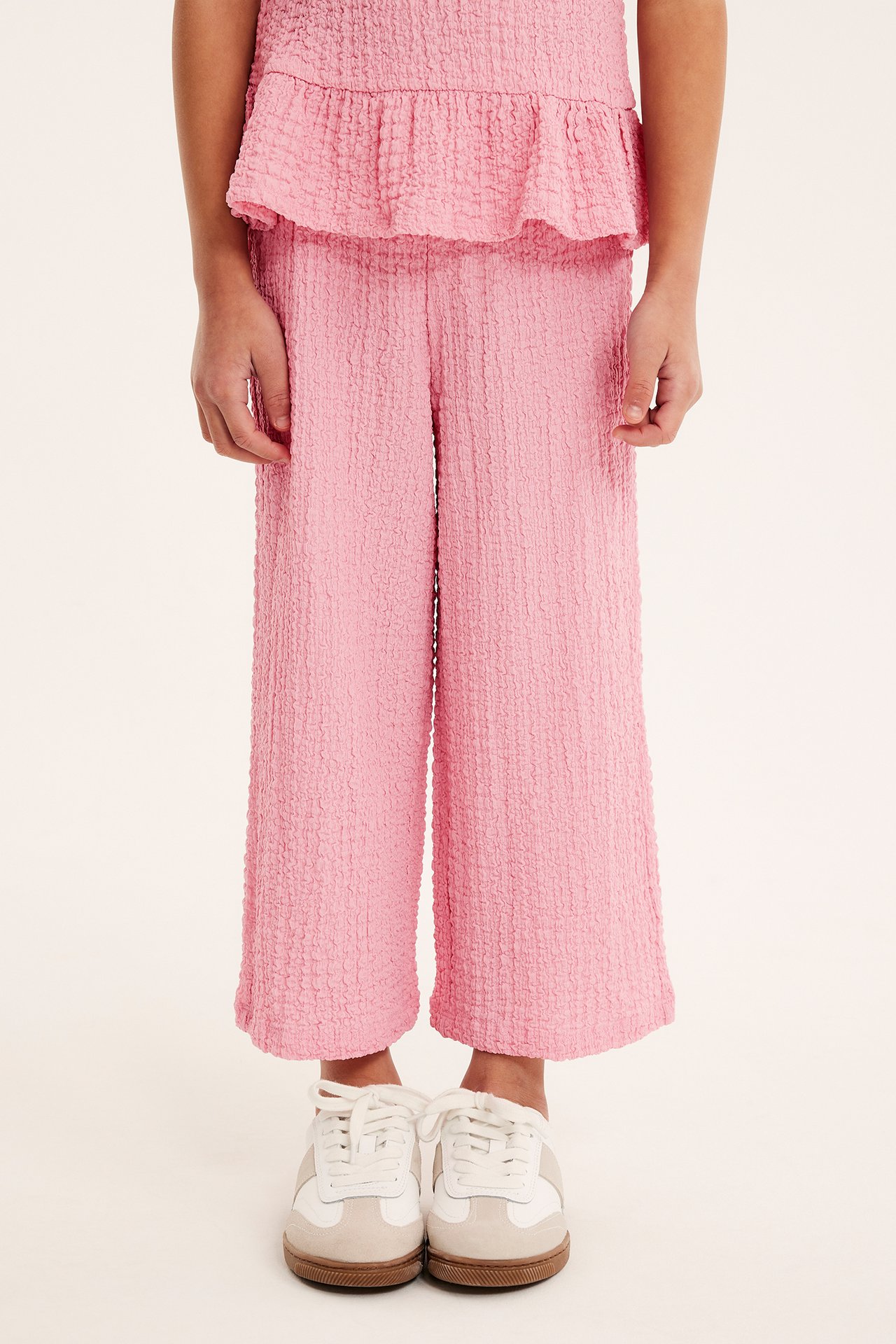 Frontvy av ett barn i rosa strukturerade culottebyxor i jersey, matchande topp och vita/beige sneakers.