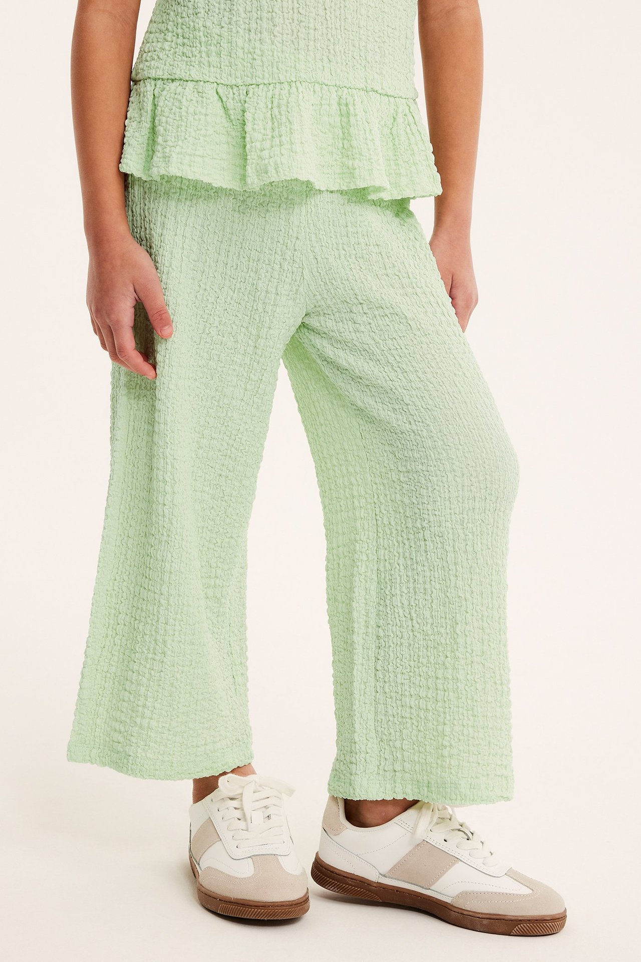 Widok z przodu dziecka w jasnozielonych, strukturalnych spodniach culotte, pasującym topie i sneakersach.