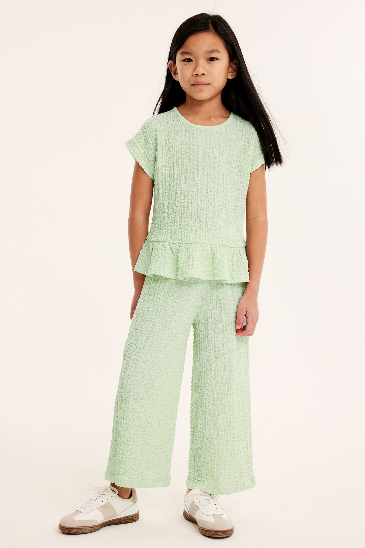Widok z przodu: dziecko w jasnozielonych, teksturowanych culotte, pasującym topie i białych sneakersach.
