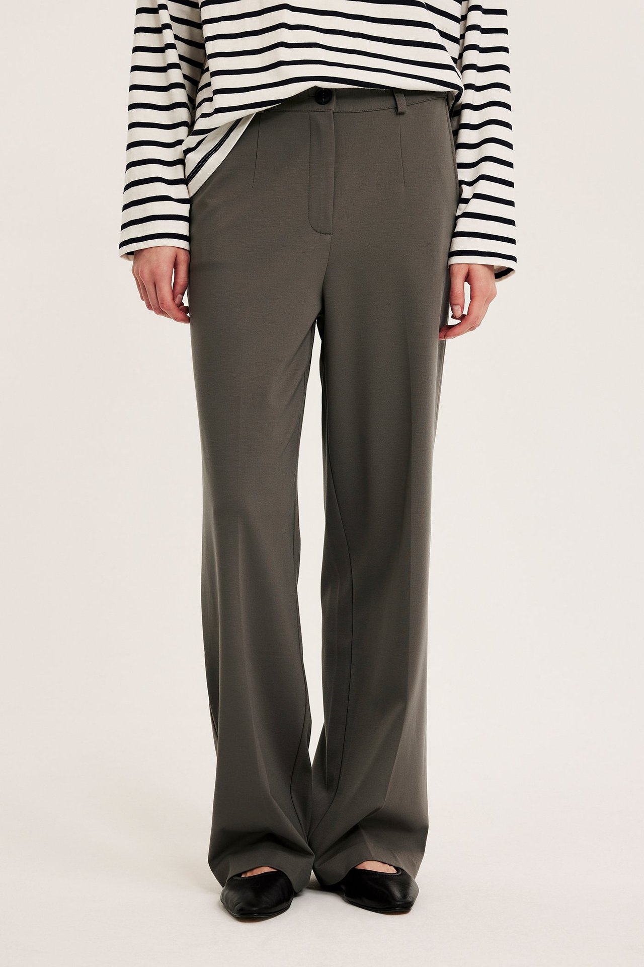 Widok z przodu kobiety w ciemnobrązowych szerokich spodniach garniturowych, bluzce w paski i czarnych balerinach.