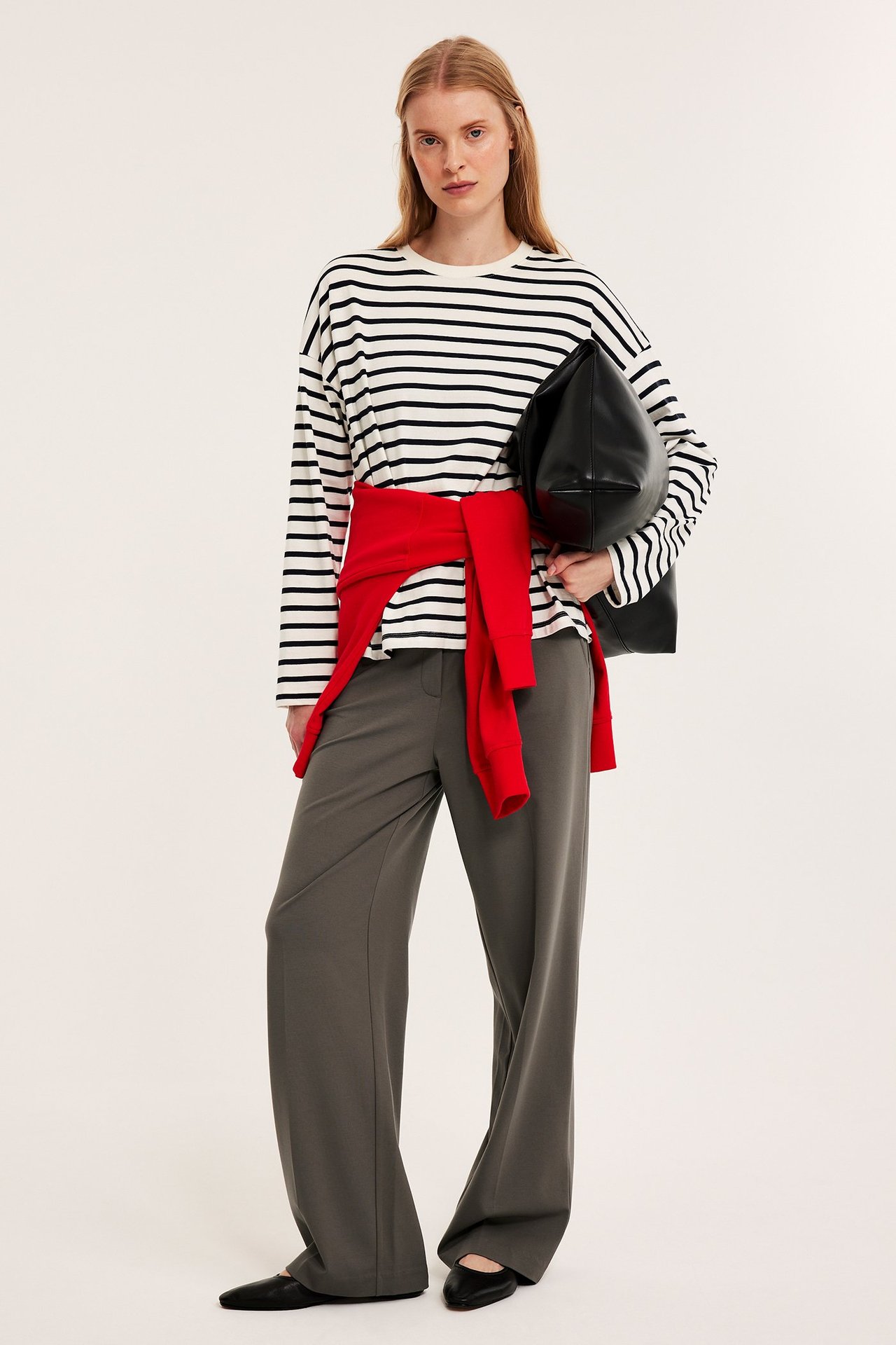 Kobieta w brązowych szerokich spodniach garniturowych, bluzce w paski, czerwonym swetrze, czarnych butach.