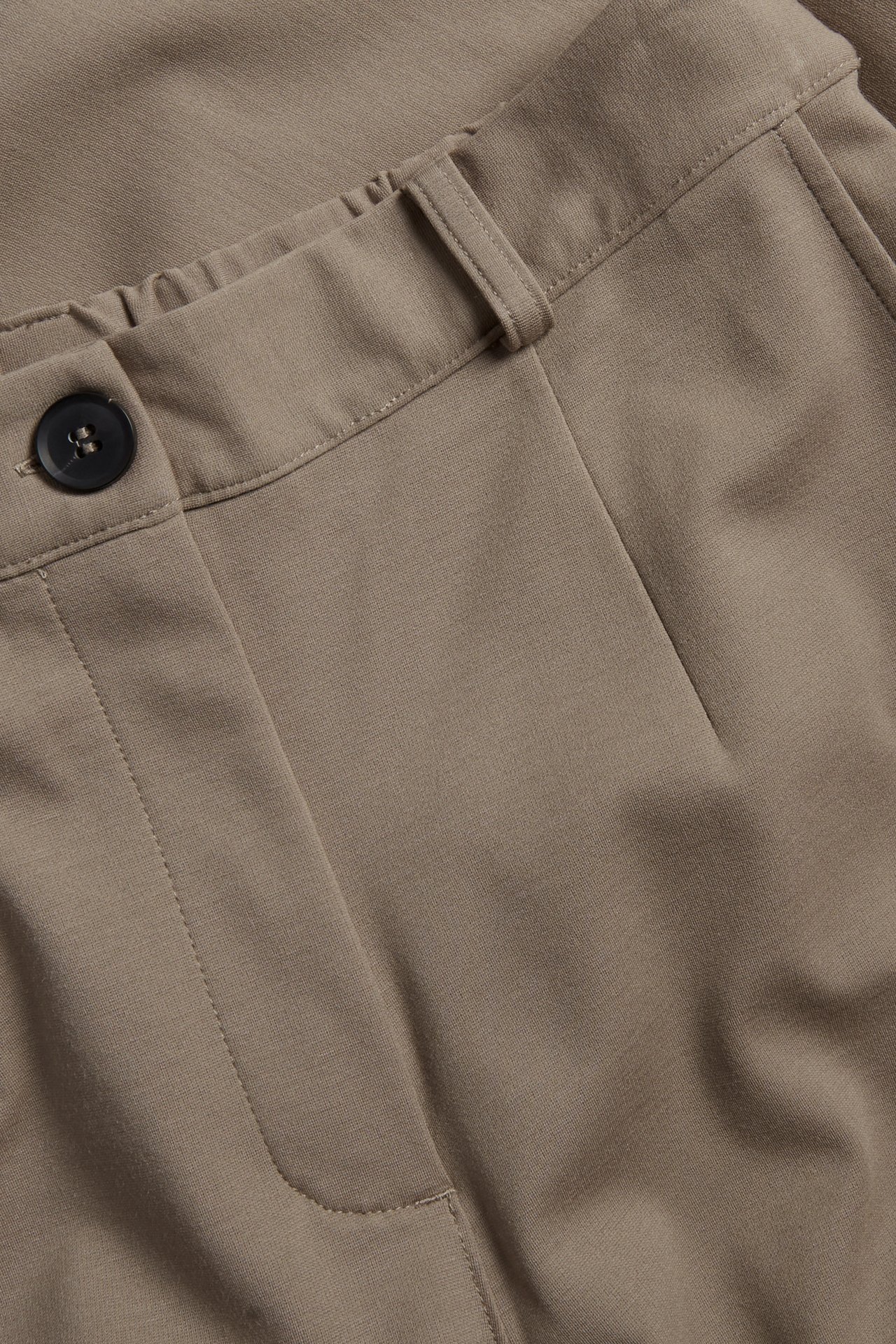 Nærbilde av mørk beige dressbukse med svart knapp og elastisk midjedetalj.