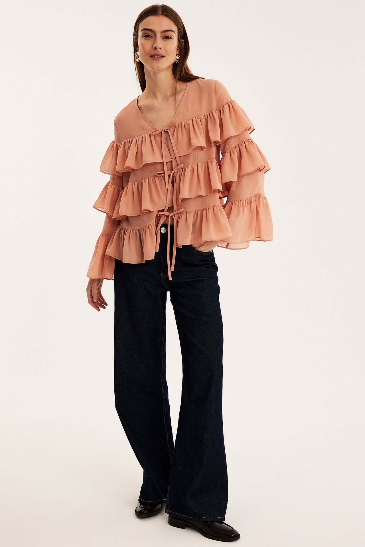 Framifrån: Kvinna bär korallfärgad volangblus med knytband, mörkblå vida jeans och svarta platta skor.