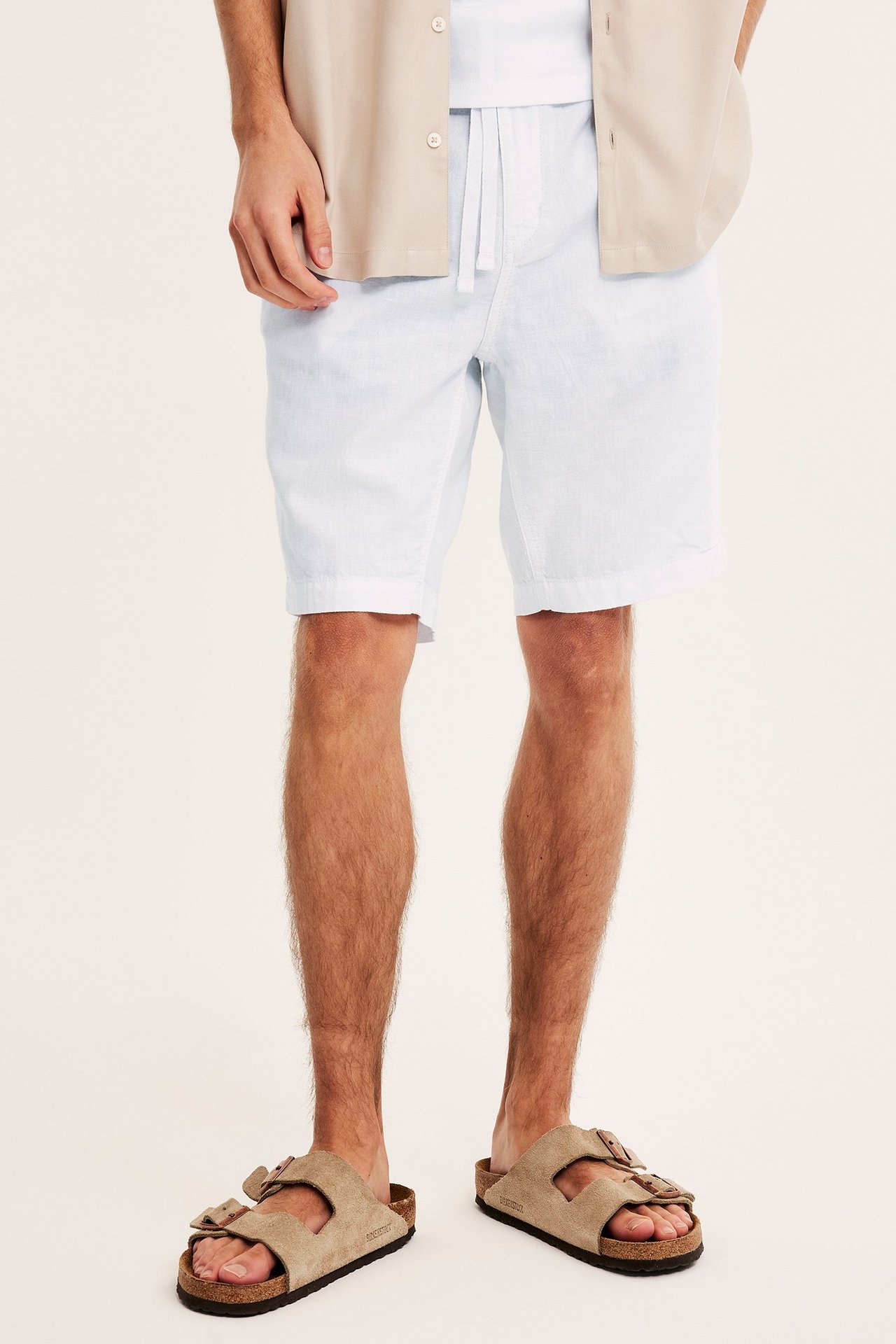 Edestä: Mies yllään valkoiset pellavasekoiteshortsit, kiristysnyöri, valkoinen t-paita, avoin beige paita ja sandaalit.