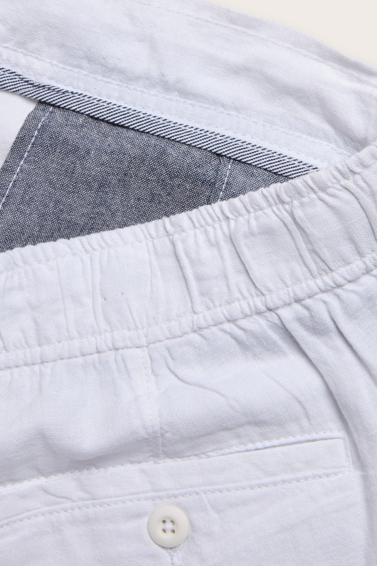 Zbliżenie białych szortów z mieszanki lnu z elastycznym pasem, białym guzikiem i pasiastą podszewką.