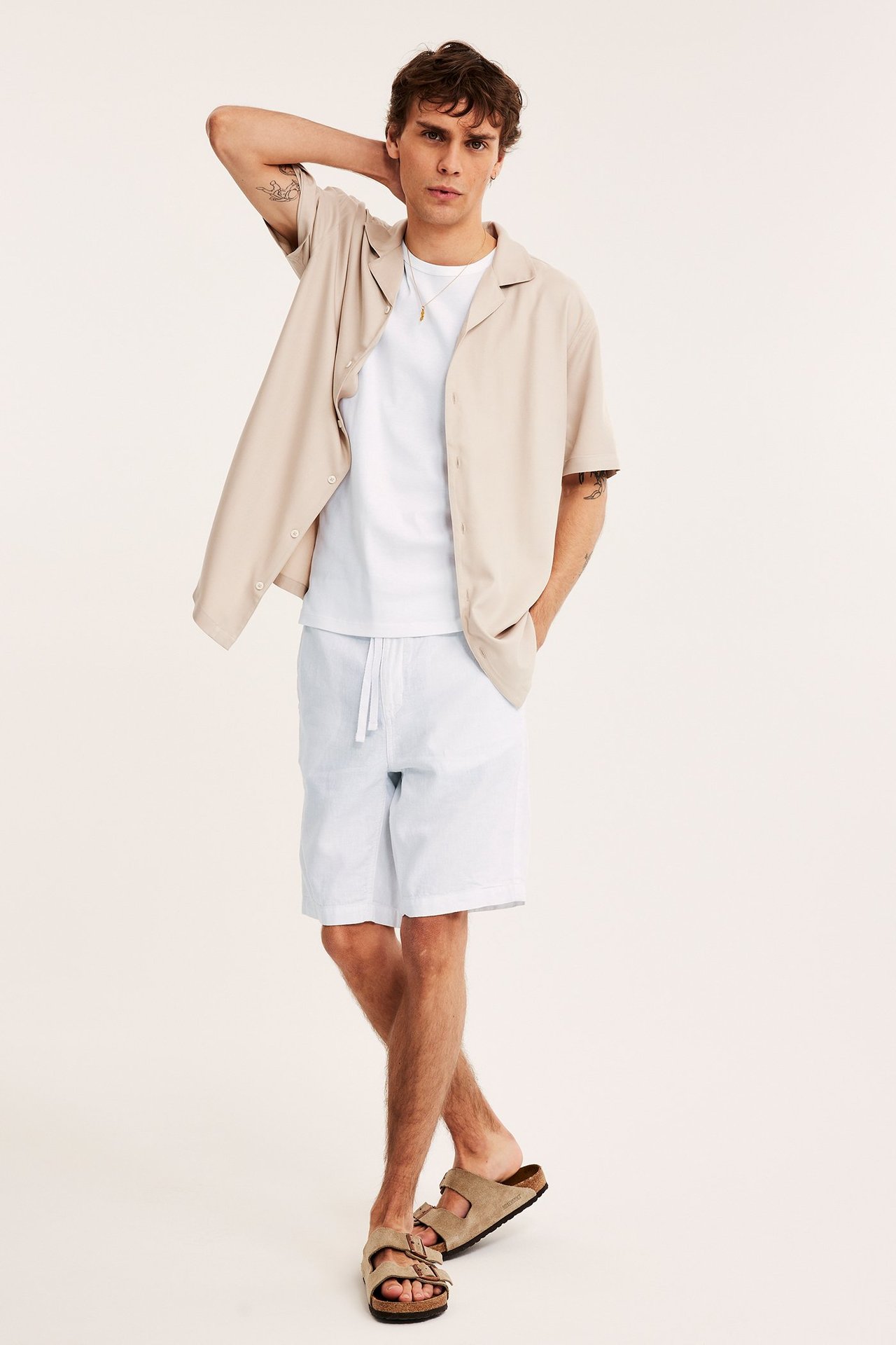 Edestä kuvattu mies vaaleansinisissä pellavasekoiteshortseissa, valkoisessa t-paidassa, beigessä paidassa ja sandaaleissa.