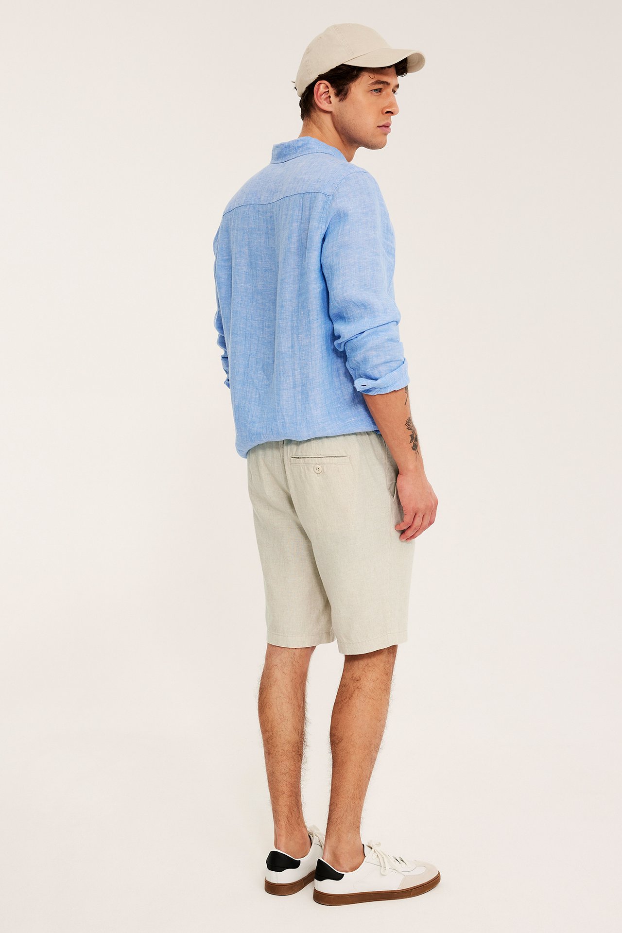 Sidevisning: mann i beige linblanding shorts, blå linskjorte, beige caps og hvite joggesko.