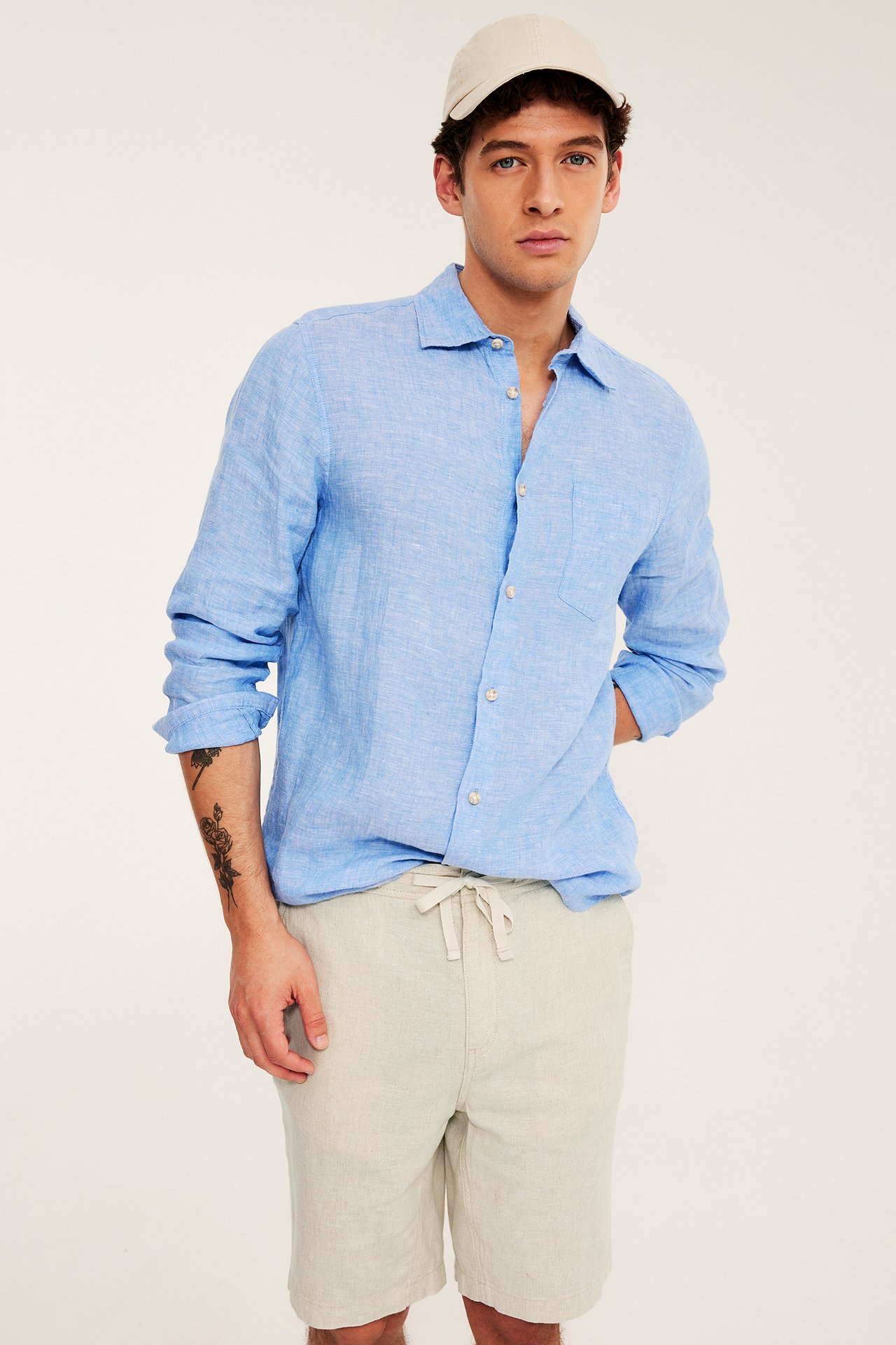 Edestäpäin mies beigessä pellavashortsissa nyörillä, sinisessä pellavapaidassa ja beigessä lippalakissa.