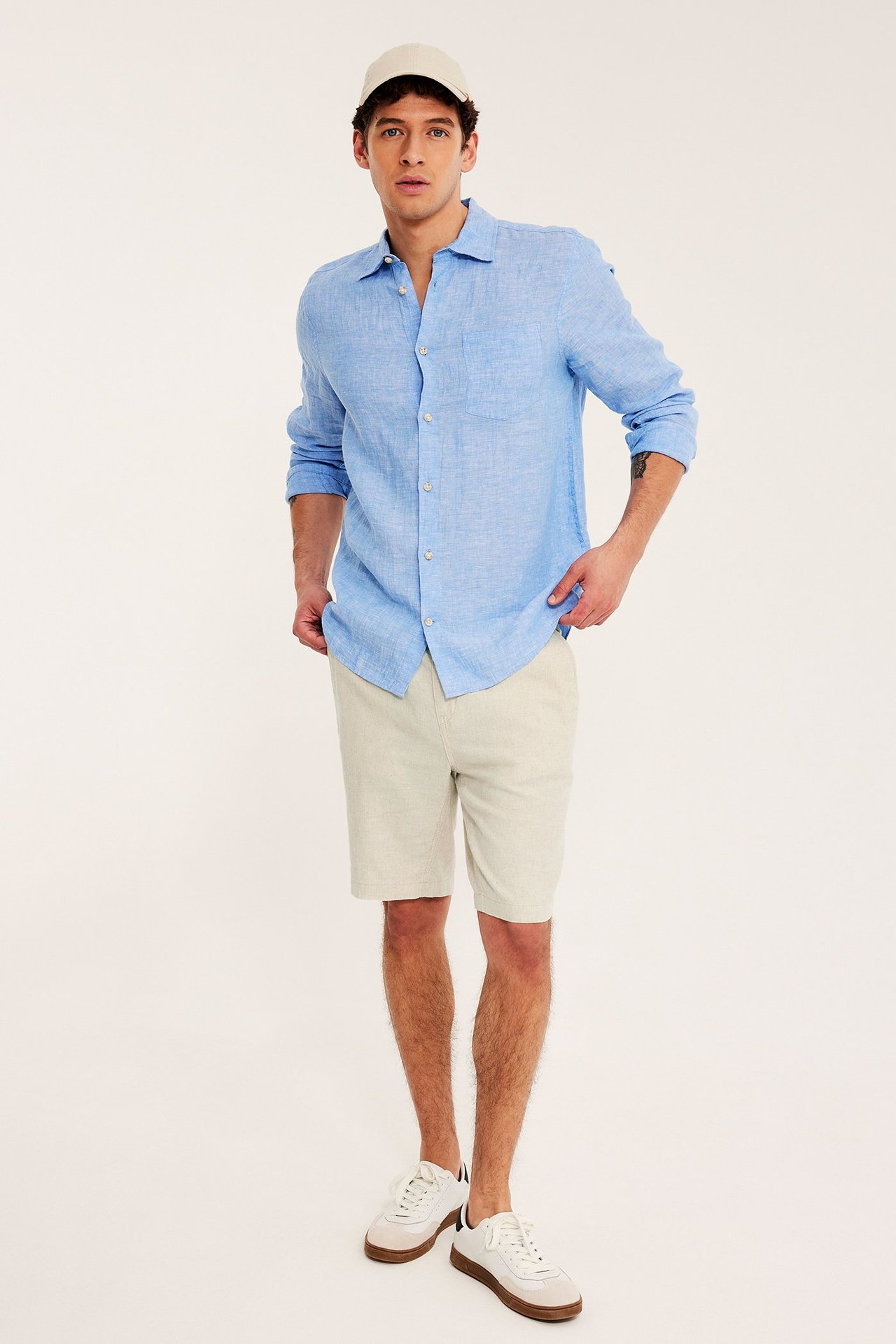 Framifrån: man i beige melange linnemixshorts med dragsko, ljusblå skjorta, beige keps och vita sneakers.