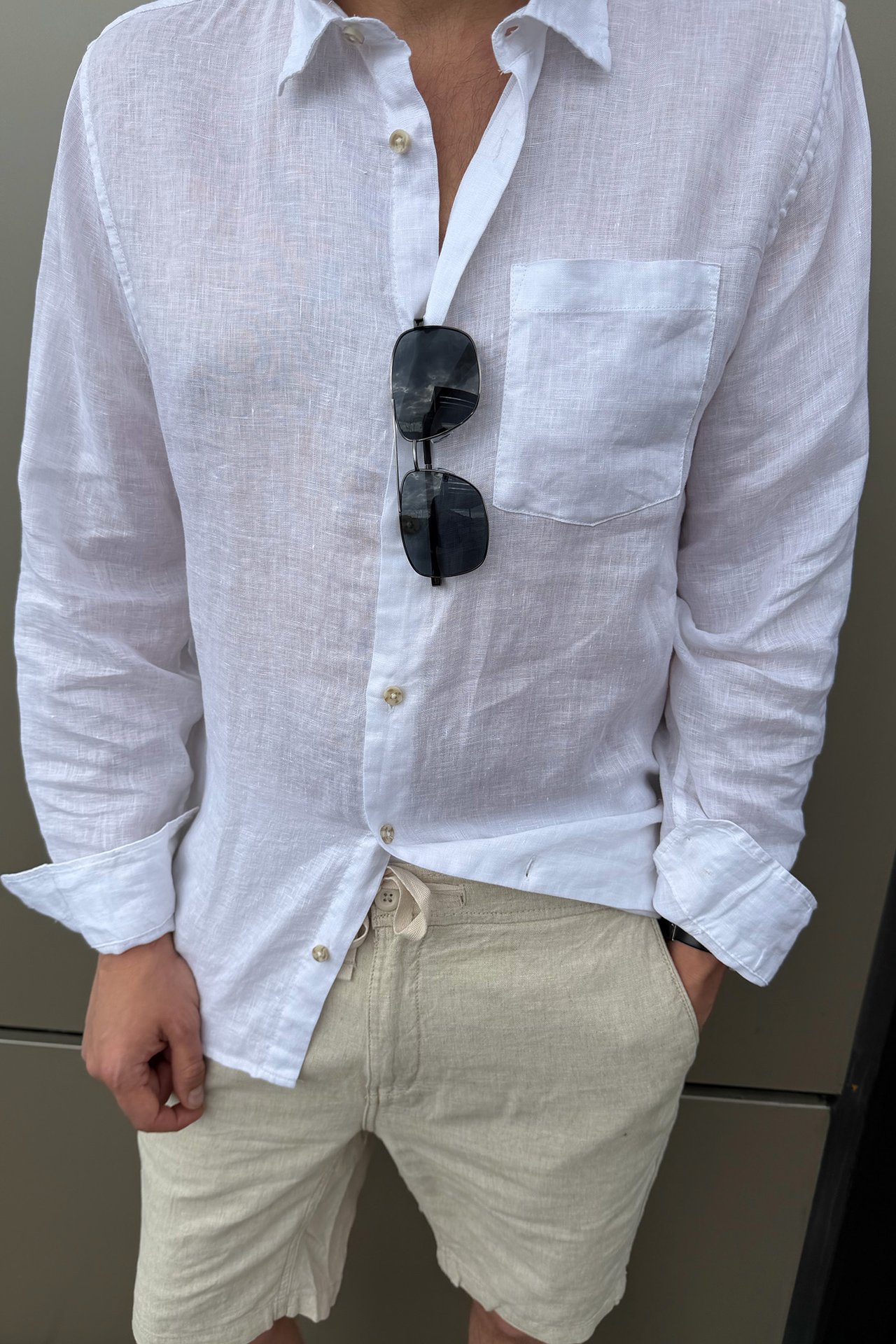 Front view: man i beiga linneshorts med dragsko, vit linneskjorta och hängande solglasögon.
