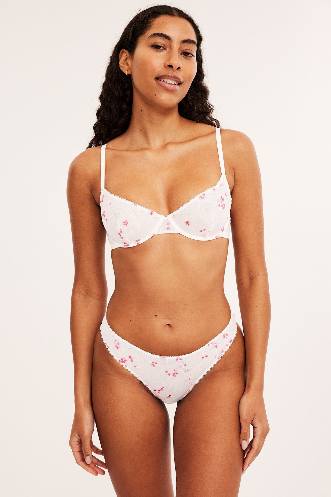 Frontvy av en kvinna i offwhite broderad bygel-bh i mesh med rosa blommor och matchande trosa.